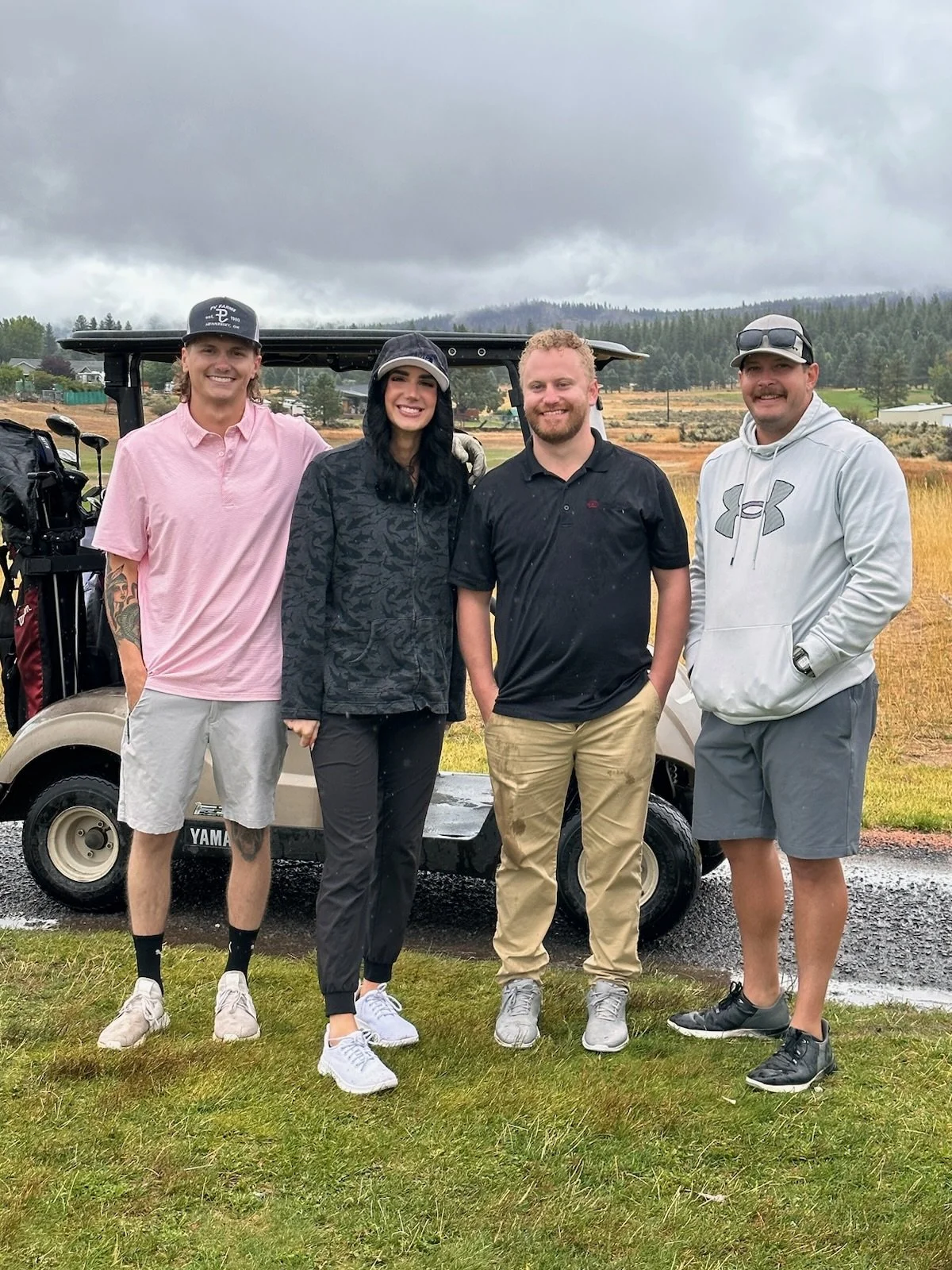 Four people standing on grass in front of a golf cart on a cloudy day, with a field and trees in the background.
