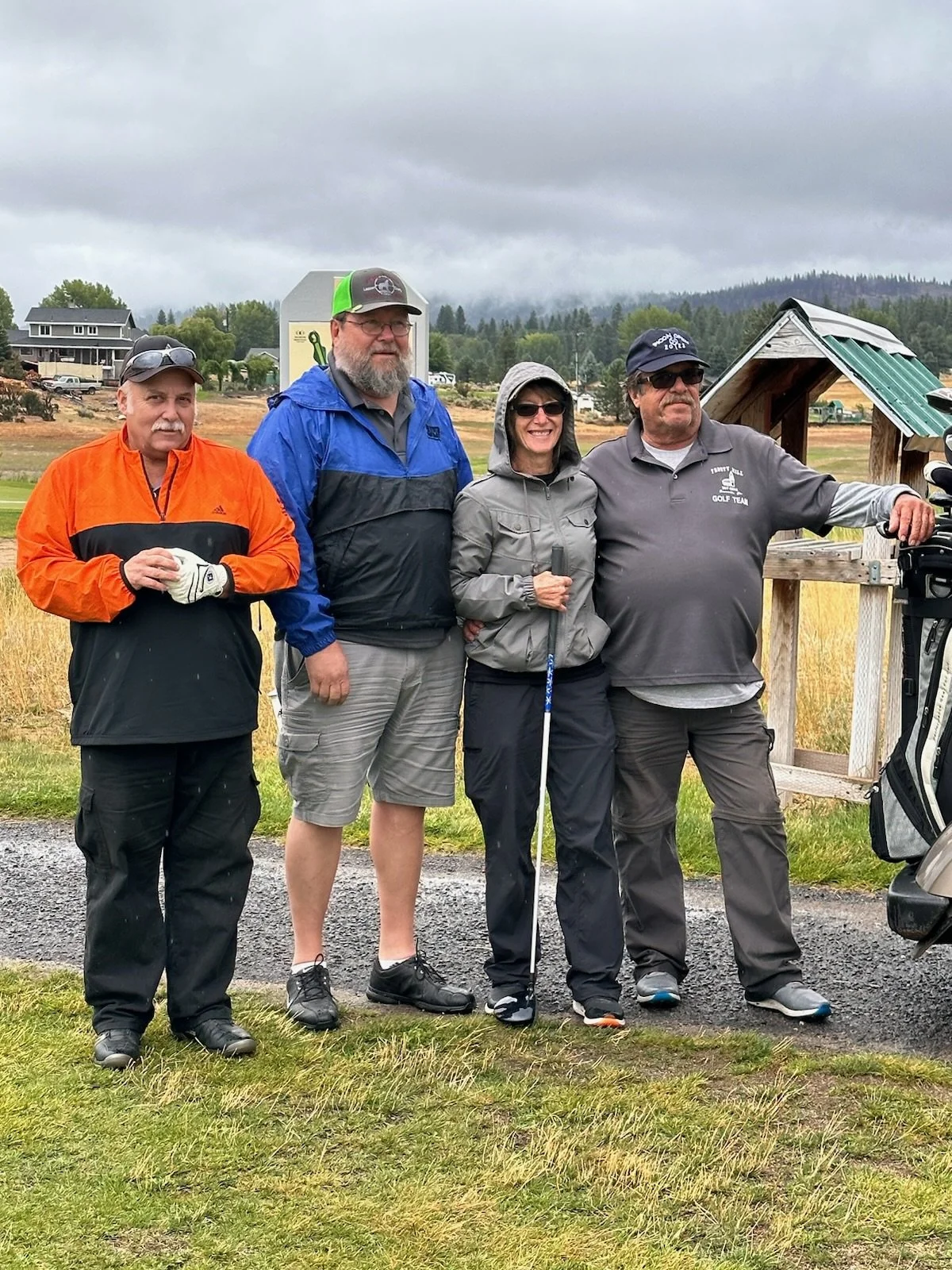 Group of four people standing on a golf course, dressed in golf attire, with a golf cart and landscape in the background under cloudy sky.