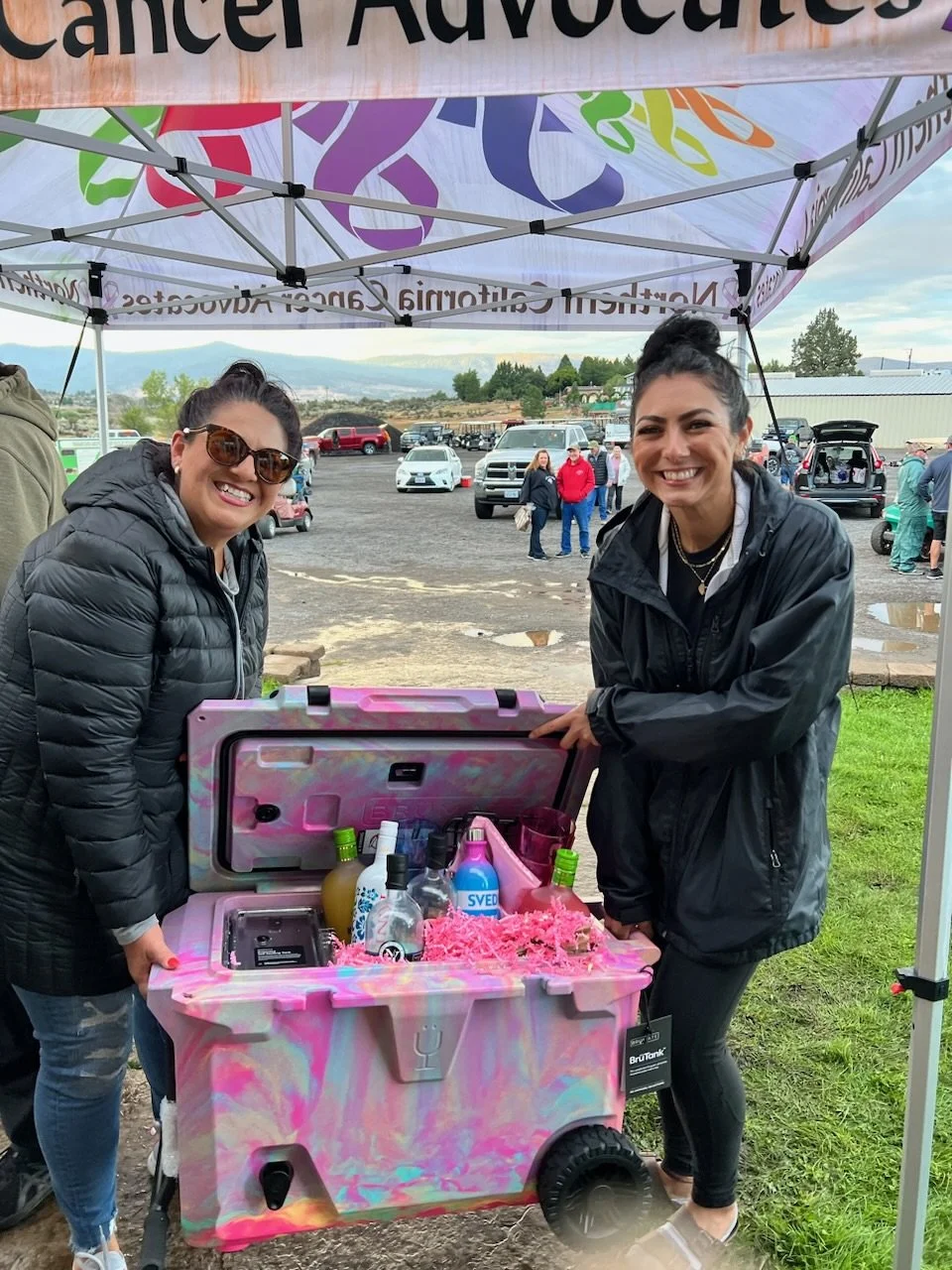 Two women standing behind a colorful cooler filled with drinks and party supplies under a canopy at an outdoor event, with parked cars and a cloudy sky in the background.
