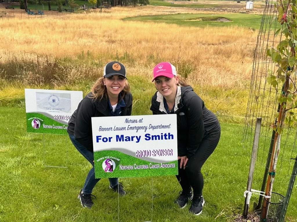 Two women in outdoor clothing crouching next to a sign supporting Mary Smith for Northern California Cancer Advocates, with a grassy field and some trees in the background.