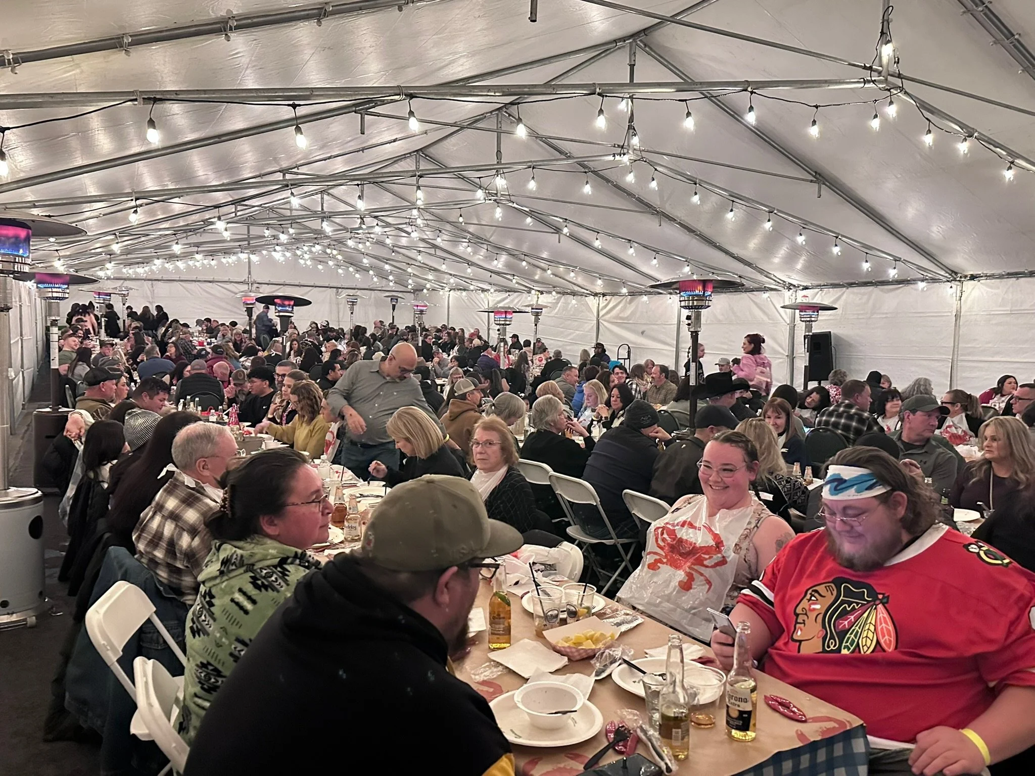 People gathered under a large tent with string lights, enjoying a meal, with heaters and tables filled with food and drinks.