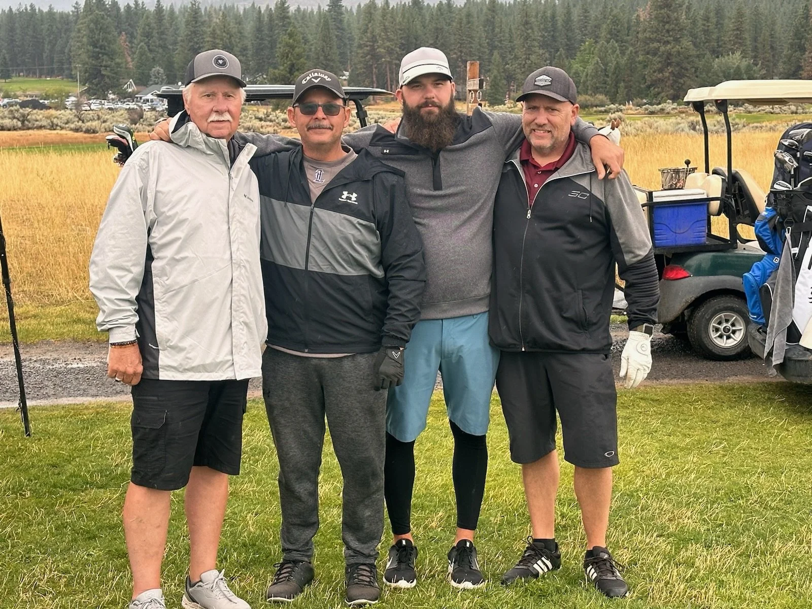 Four men standing together on a golf course, smiling, with golf carts and greenery in the background.