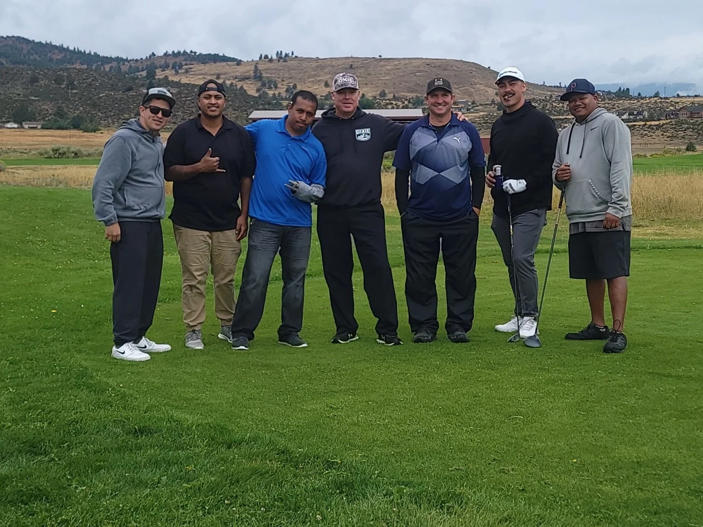 Seven men standing together on a golf course, posing for the photo with a scenic landscape of hills and fields in the background.