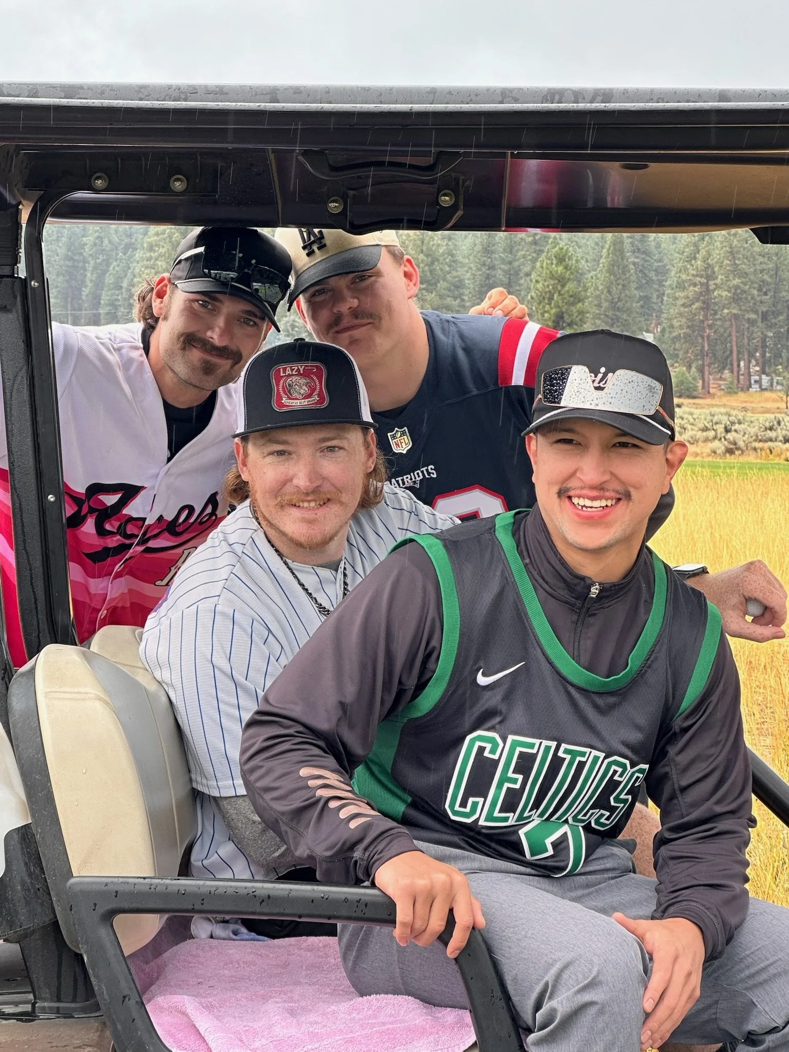 Four young men sitting in a golf cart outdoors in a grassy, wooded area, smiling at the camera.