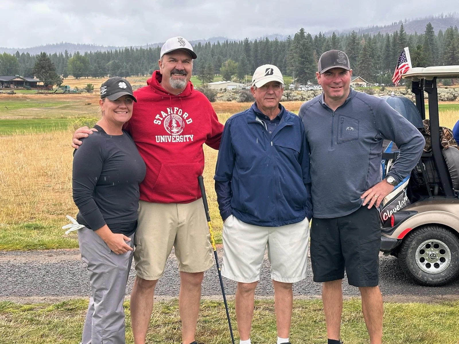 Four people standing on a golf course with a golf cart, lush trees, and mountains in the background. They are dressed in golf attire and smiling.