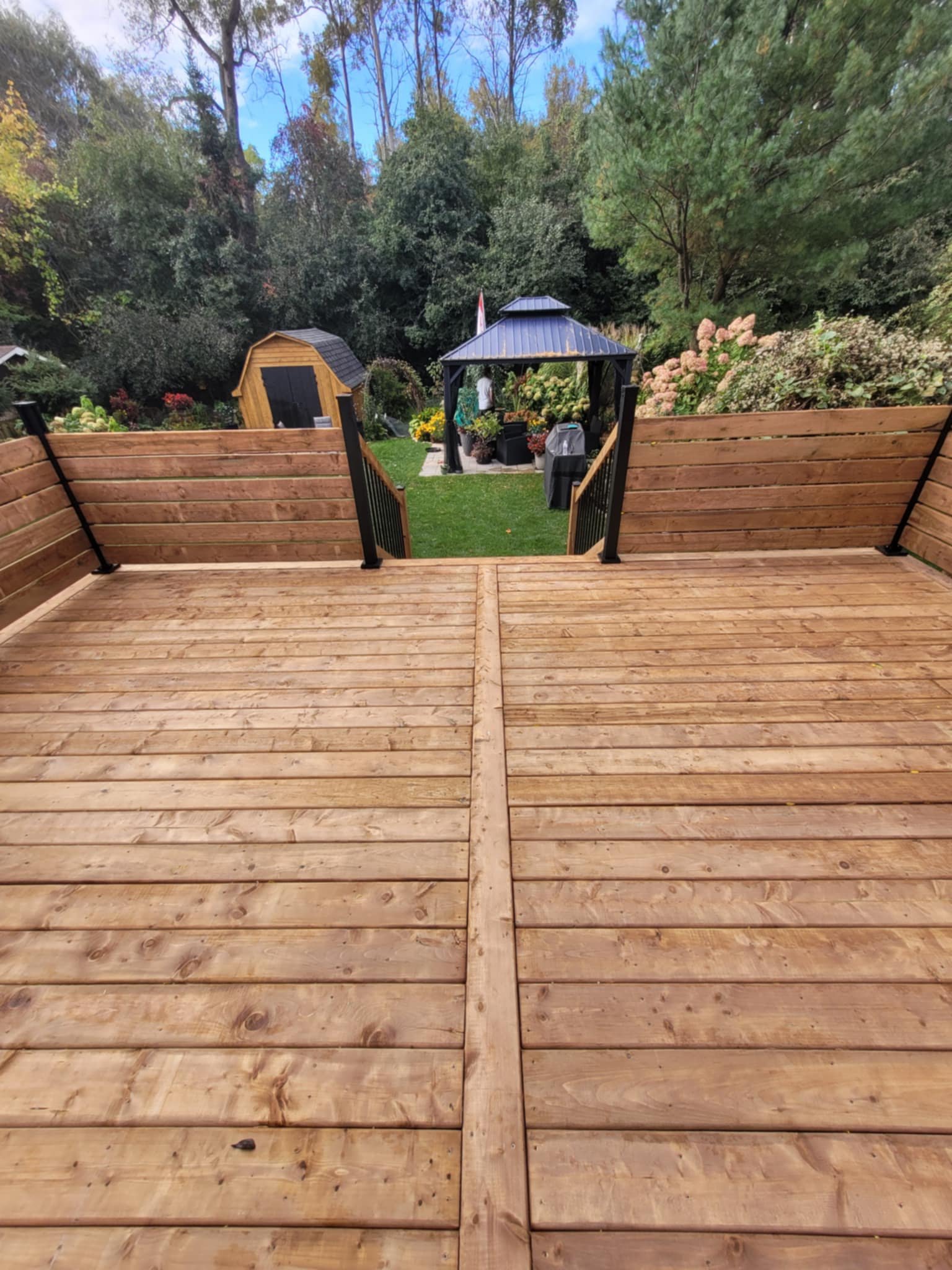 A view from a wooden deck looking out over a grassy backyard with a pavilion, garden shed, and various plants, trees, and shrubs in the background.