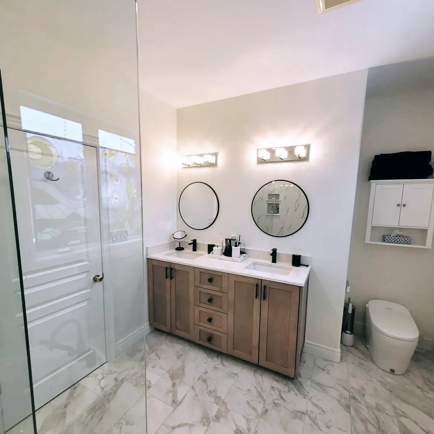 Modern bathroom with a dual sink vanity, round mirrors, black fixtures, marble floor, glass shower door, toilet, and storage cabinet.