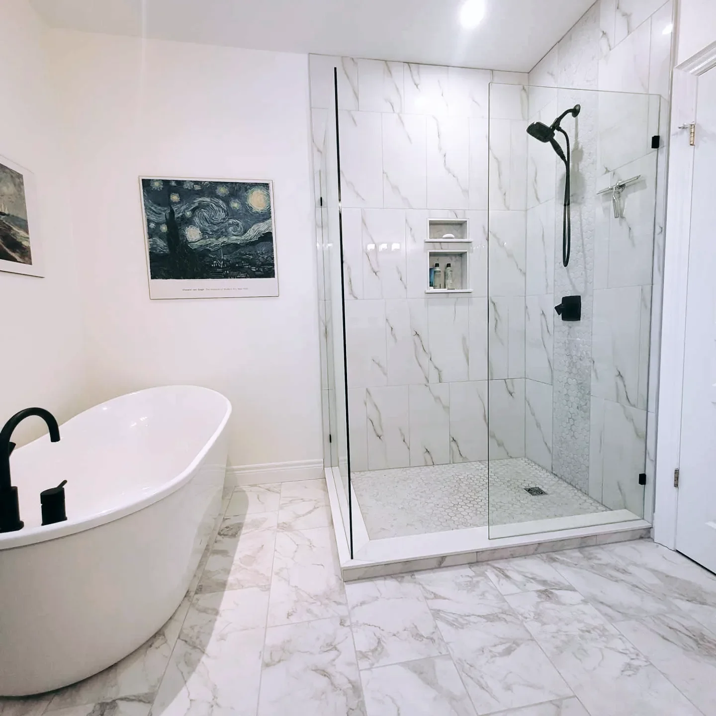 Modern bathroom with white marble floor and walls, a white freestanding bathtub with black fixtures, and a glass-enclosed walk-in shower with marble tiles, built-in shelves, and a black showerhead.
