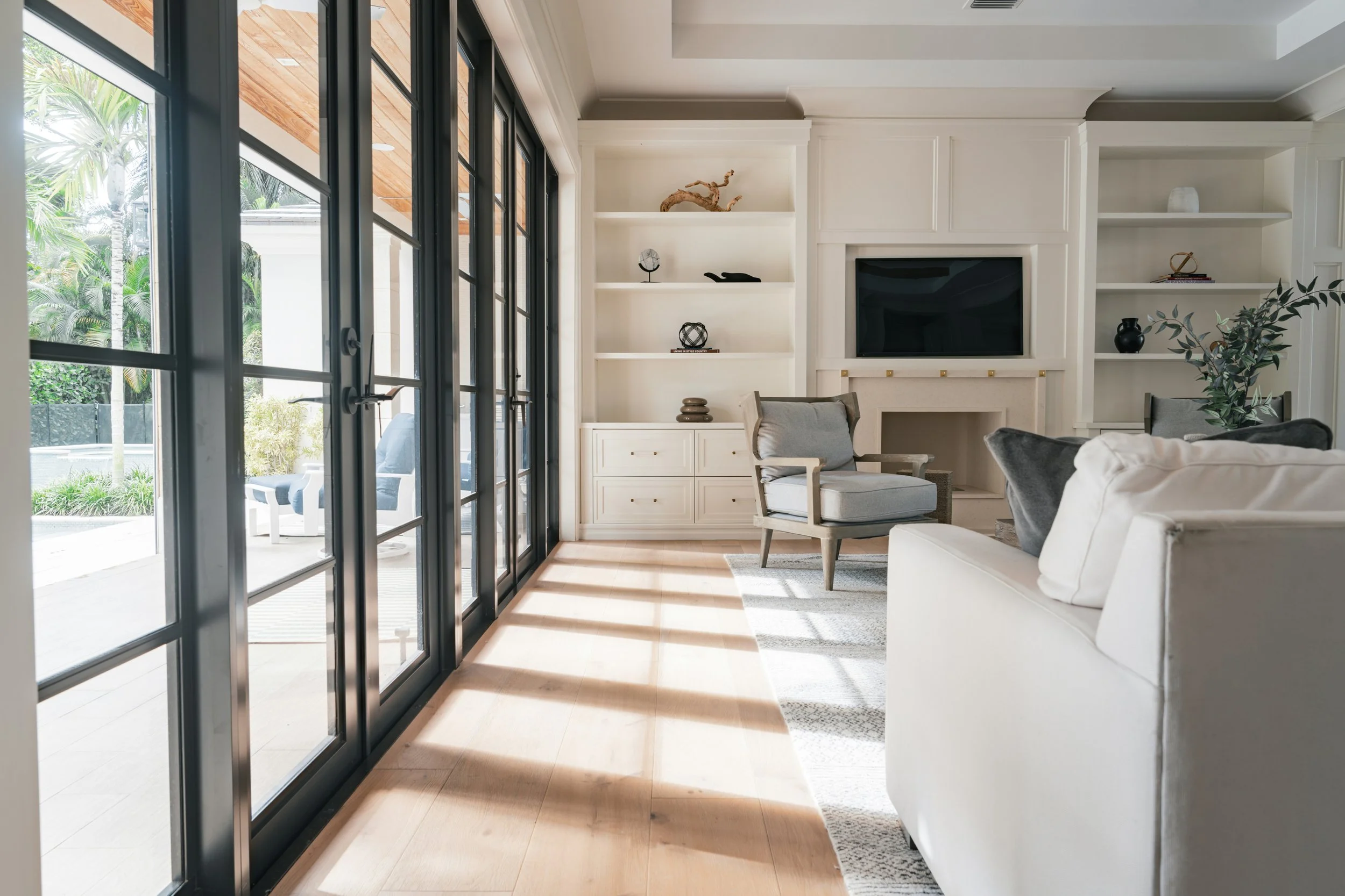 Interior of a living room with large glass sliding doors leading outside, white built-in shelves with decorative objects, a black flat-screen TV, a white armchair with a pillow, and a beige sofa. Outside, patio furniture and lush greenery are visible.