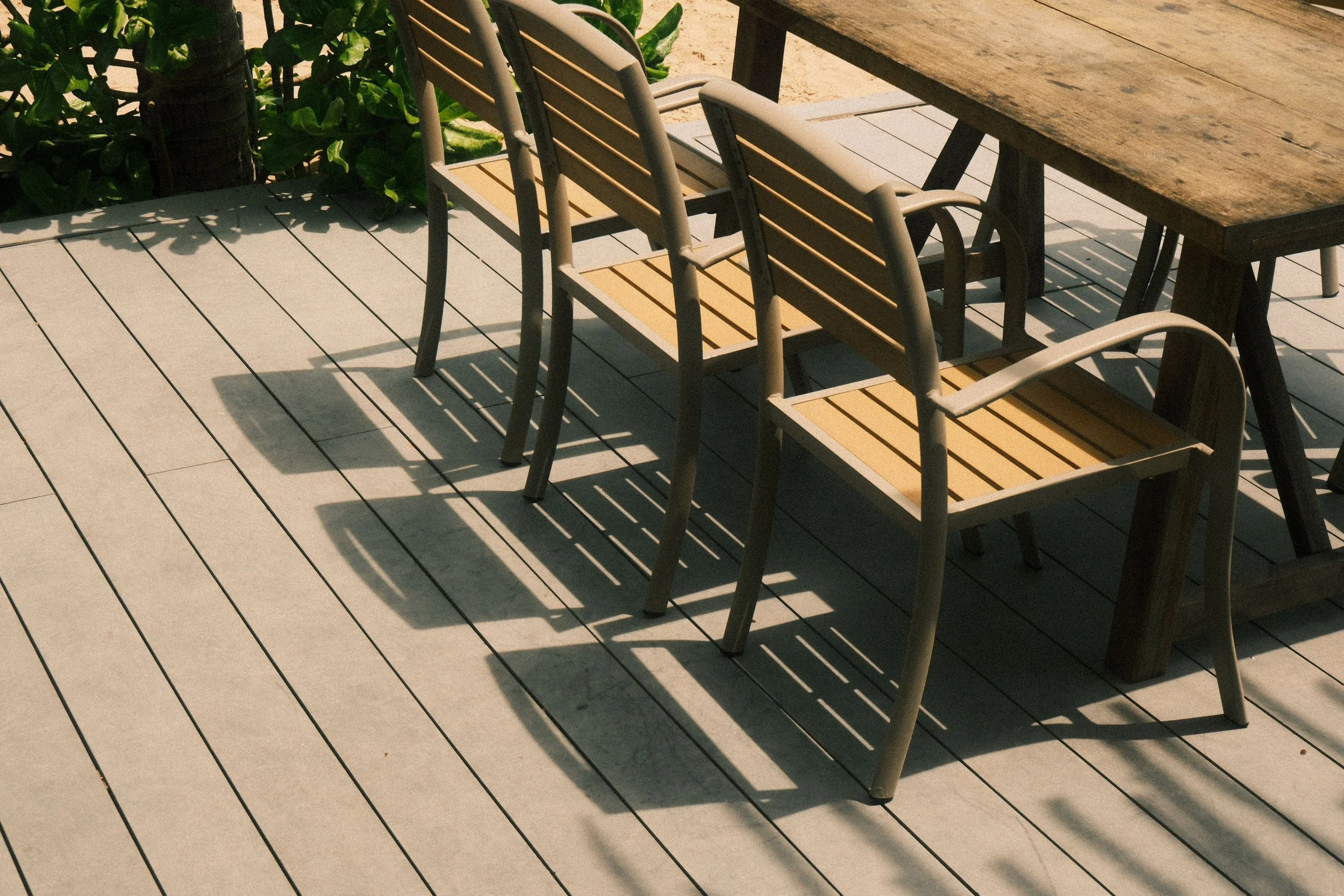 Empty wooden and metal chairs around a wooden table on a deck with shadows cast by the chairs.