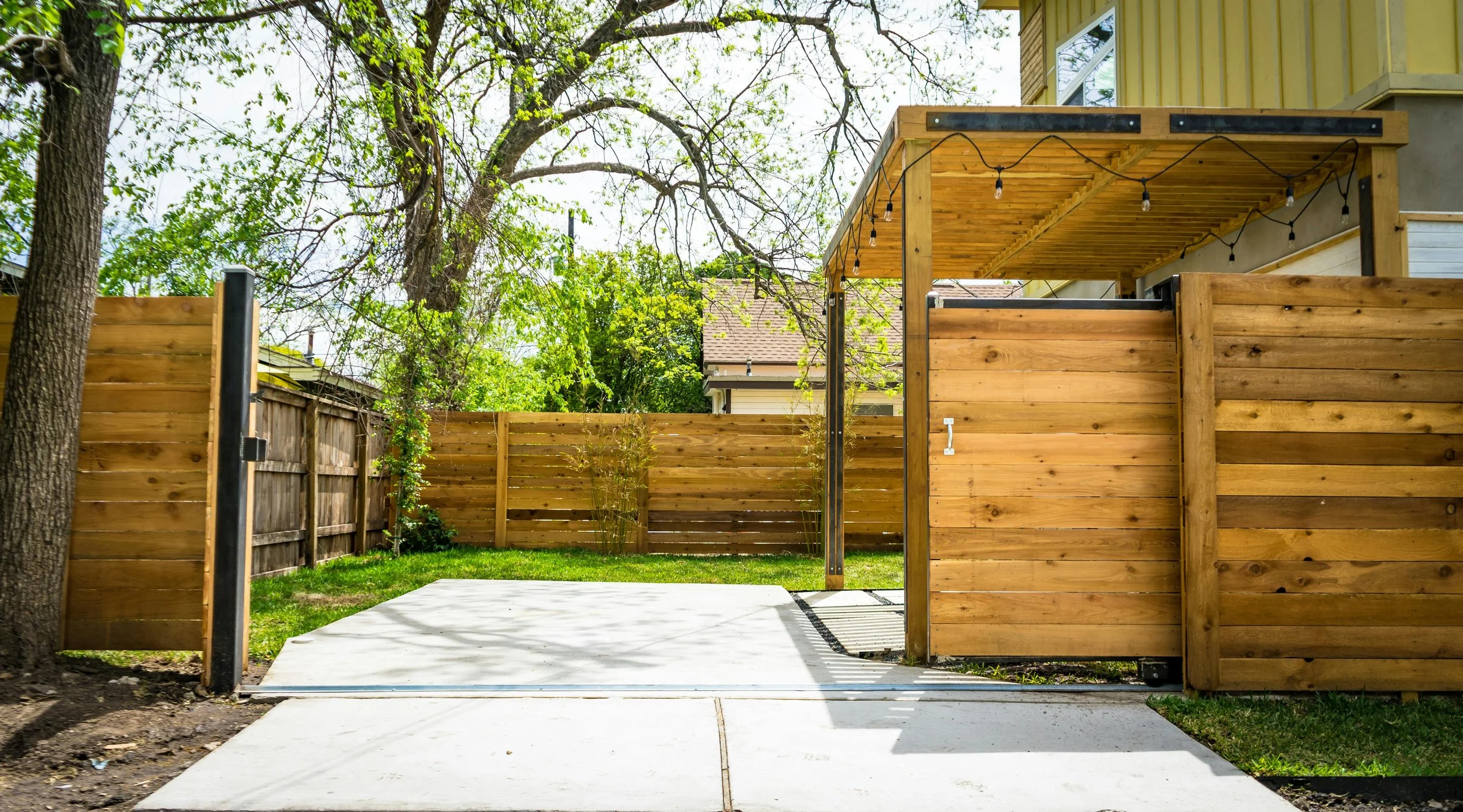 A backyard enclosed by a wooden fence with gate, featuring a tree with branches extending over the fence, a concrete patio, and a new wooden pergola draped with string lights.