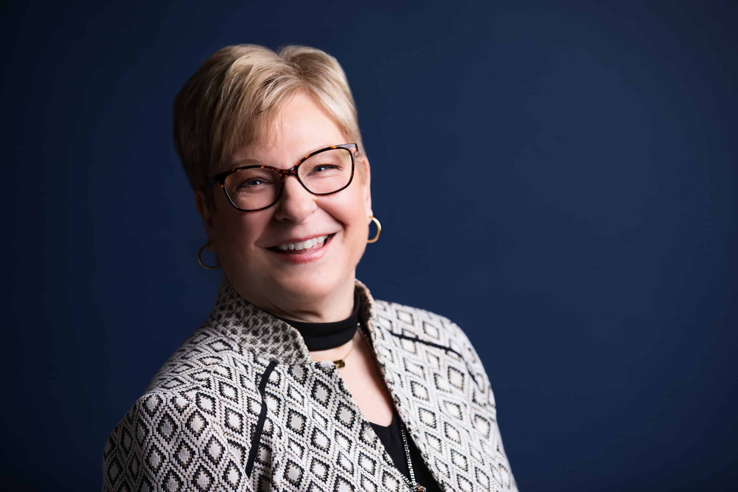A smiling woman with short blonde hair and glasses, wearing a patterned blazer and earrings, against a dark blue background.