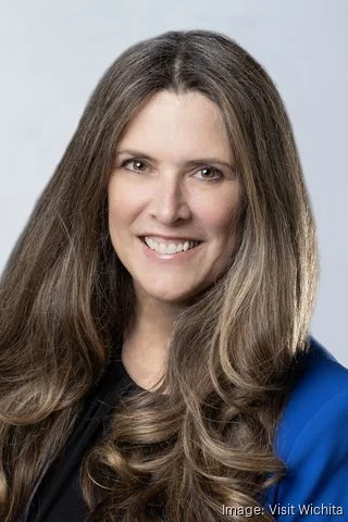 Close-up portrait of a woman with long, wavy brown hair, smiling, wearing a black top and a blue blazer.