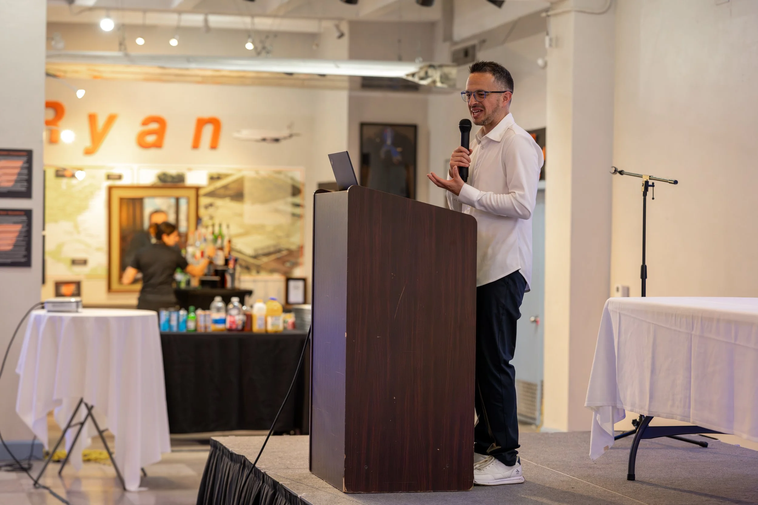 A man in a white shirt and dark pants is giving a presentation on a small stage, speaking into a microphone. There is a laptop on the podium. Behind him, a bar area and a woman preparing drinks are visible, with a blurred sign that spells 'Bryan' on the wall.