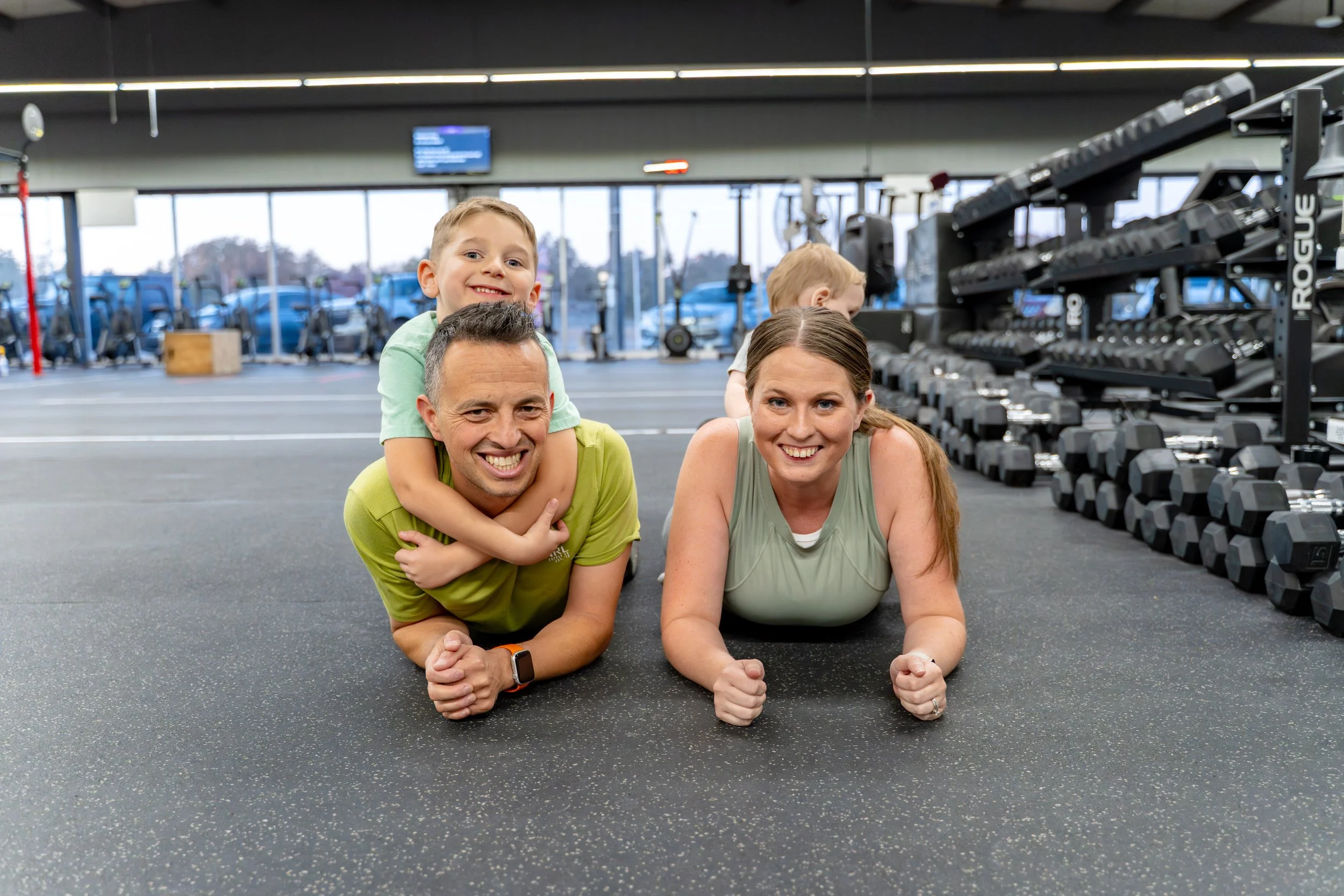 A family of four doing planks on the gym floor, with a man and woman in the foreground and two children, one on the man's back and another in the background, at a gym with poles and weights.