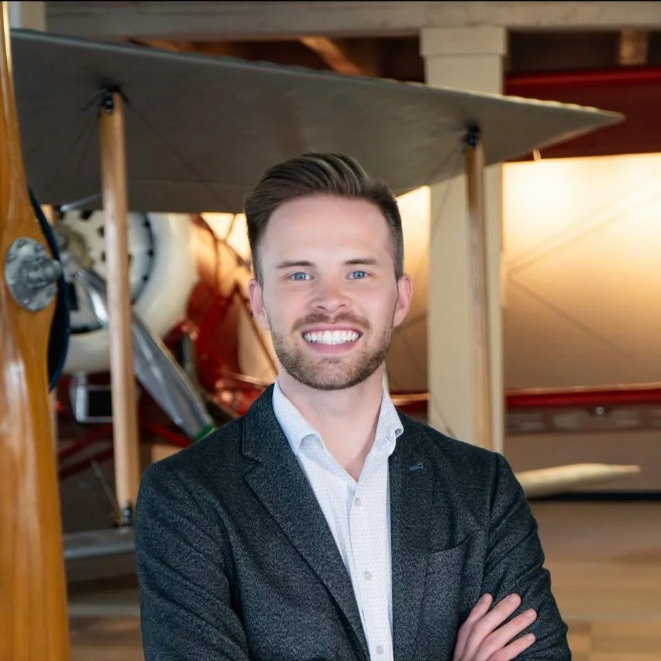 A smiling man with short brown hair, blue eyes, and a beard, wearing a white shirt and dark blazer, standing with arms crossed indoors with wooden structures and an airplane in the background.