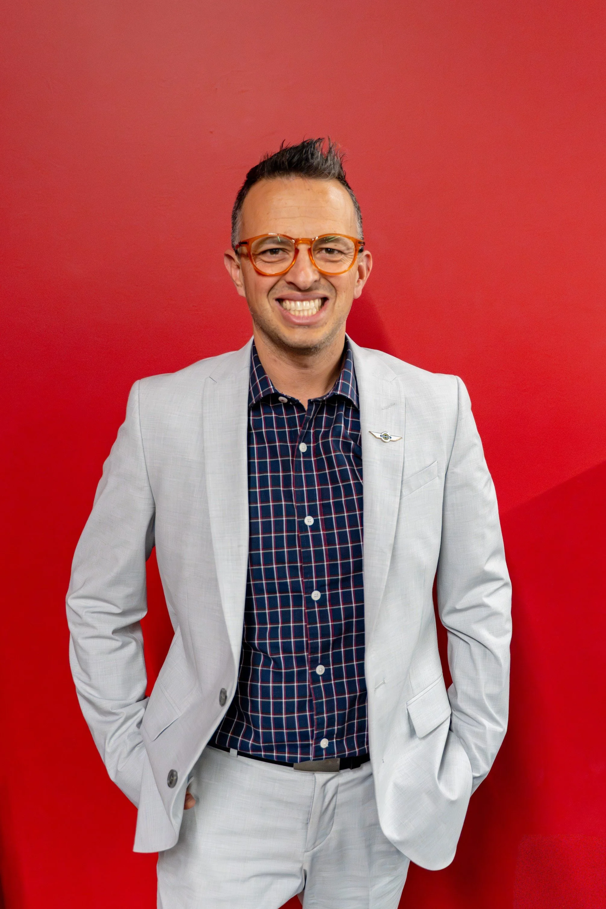 A smiling man in a light gray suit with a checkered shirt and orange glasses standing in front of a red background.