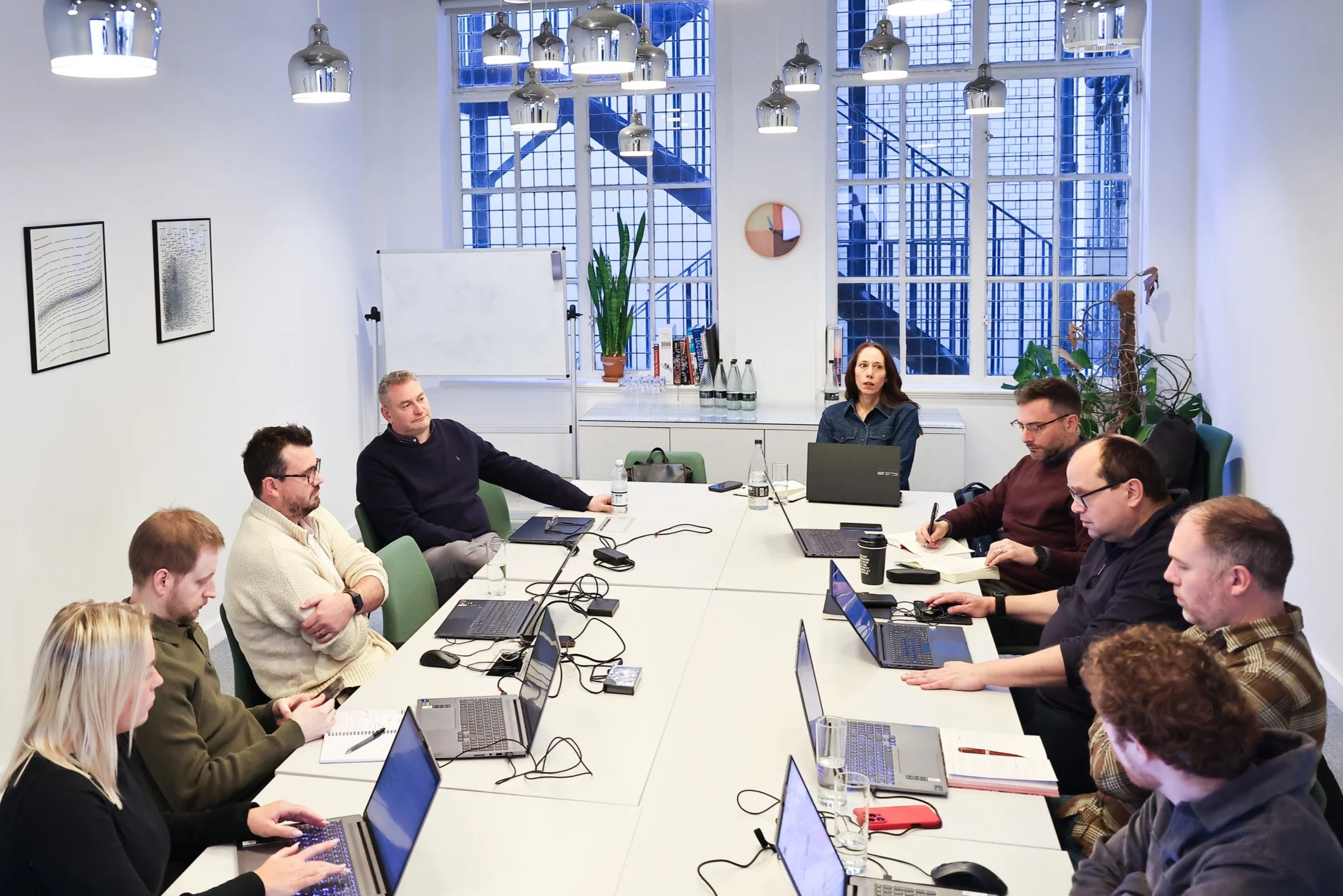 A diverse group of ten people in a modern, well-lit conference room with laptops, notebooks, and water bottles on the table, engaged in a meeting. The room has large windows with a staircase visible outside and minimalistic wall art.