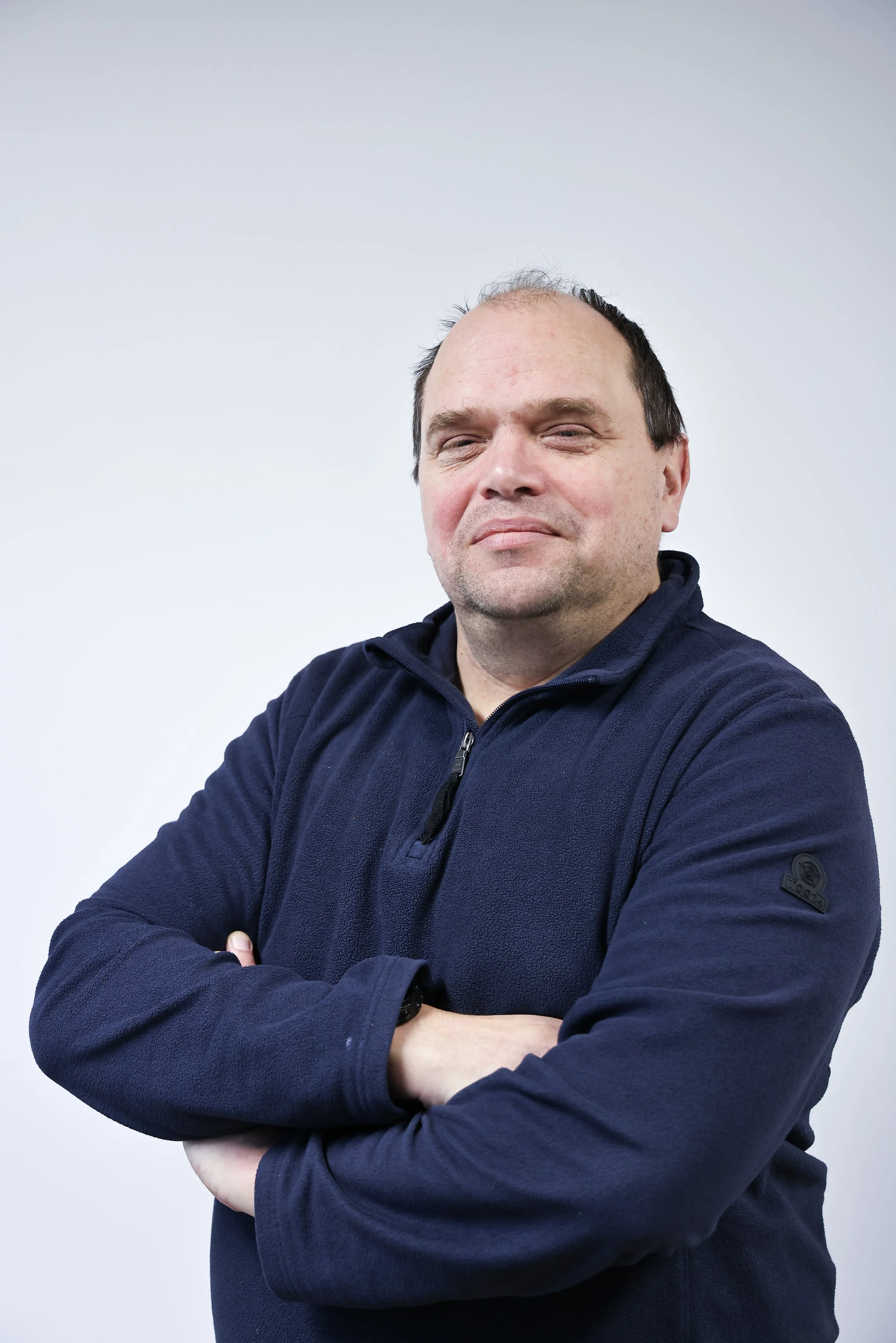 A smiling man with light brown hair and a beard, wearing a blue button-up shirt, standing against a plain gray background.