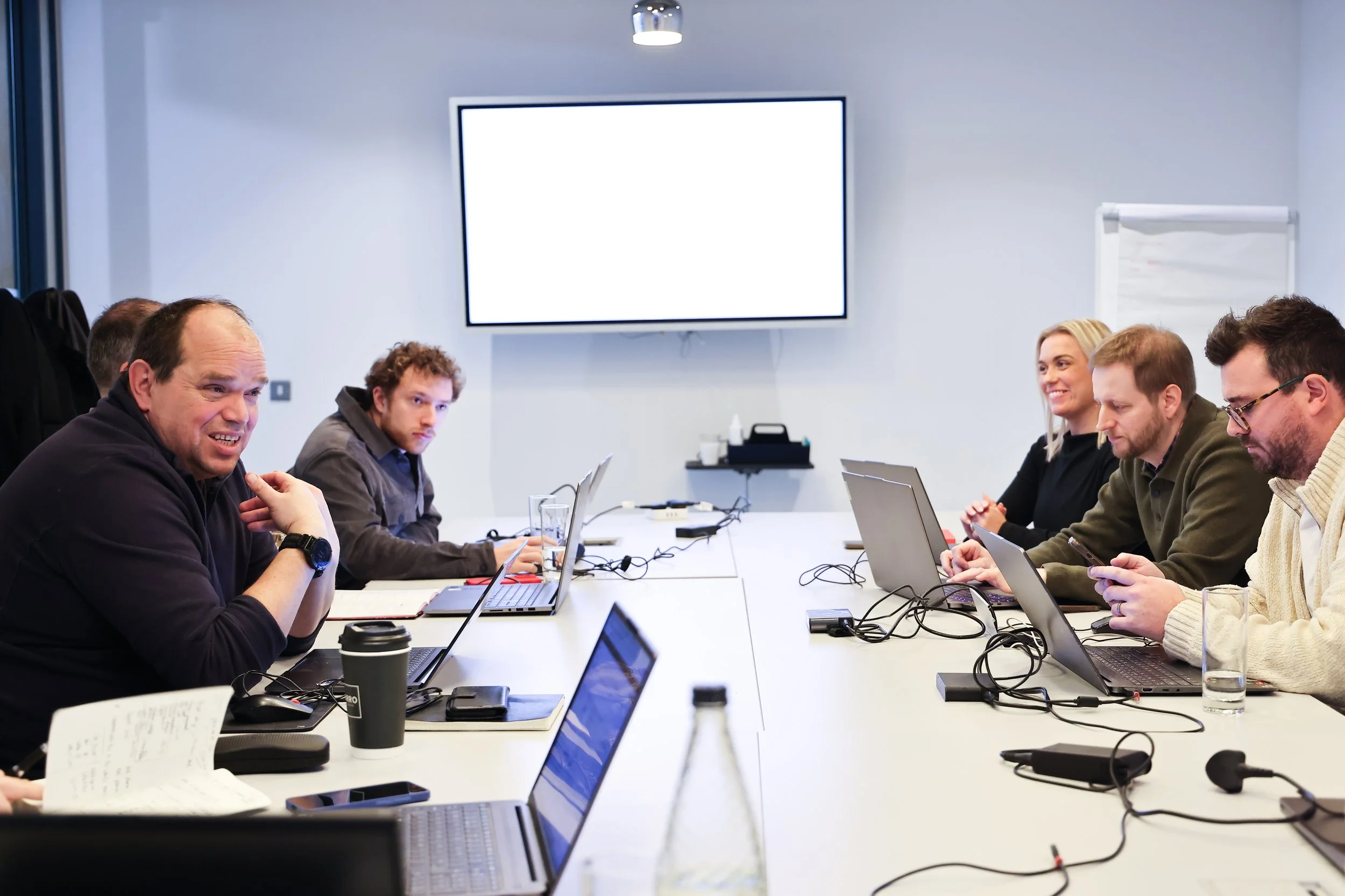 Group of people seated around a conference table engaged in a meeting, with laptops, notebooks, and drinks in front of them, in a modern office room.