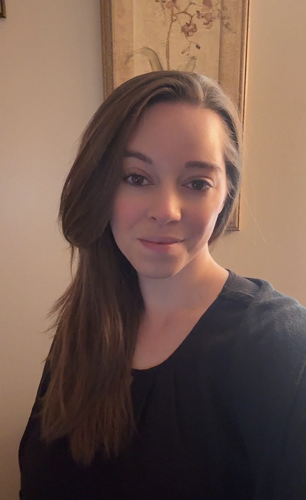 A woman with long brown hair and fair skin, wearing a black top, posing indoors in front of a wall with a framed picture.