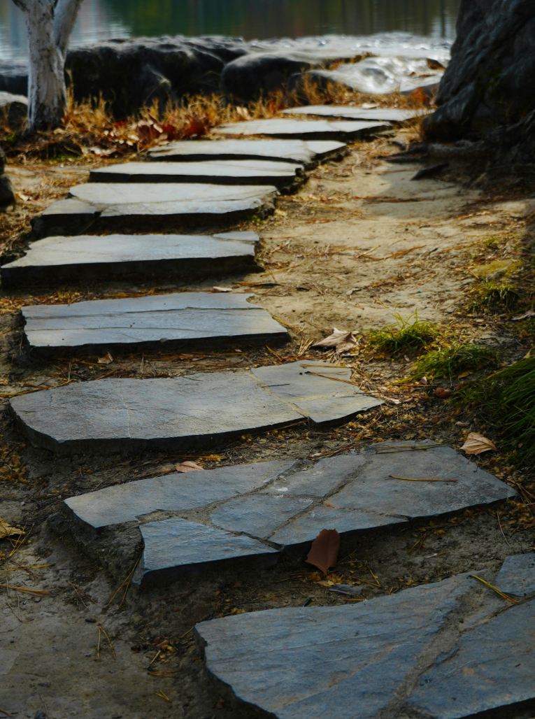 Stone pathway leading along a lakeside, with trees and rocks in the background.