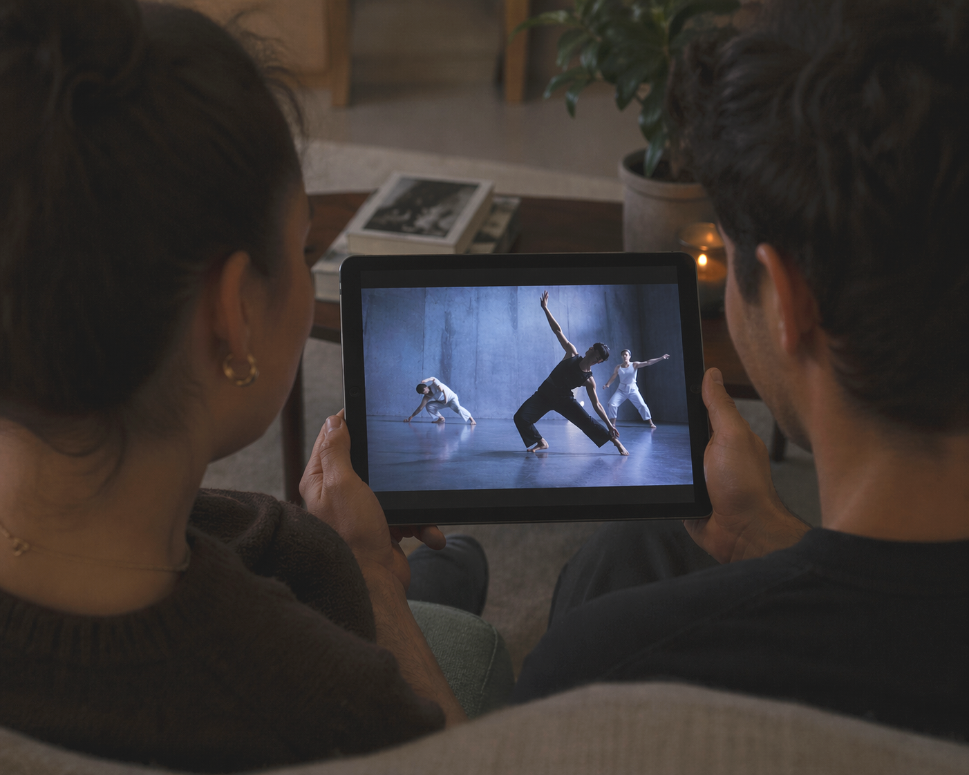 Two people sitting on a couch watching a tablet screen showing a dance practice with three dancers on stage.