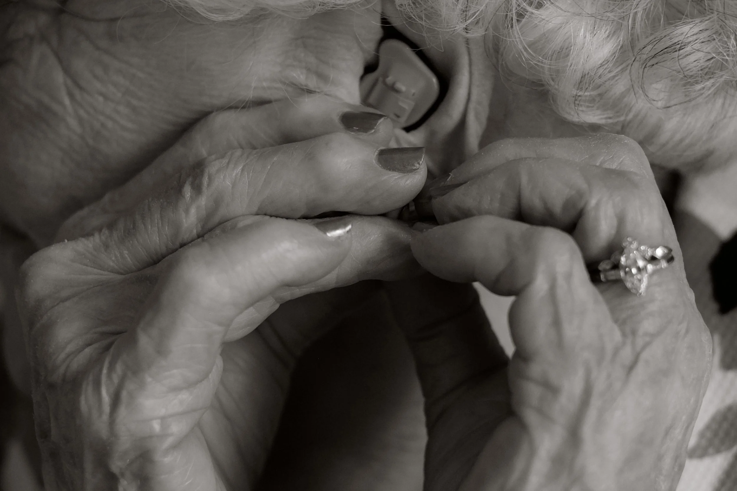 Close-up of elderly woman's hands holding a ring and touching her face, with a hearing aid visible in her ear.