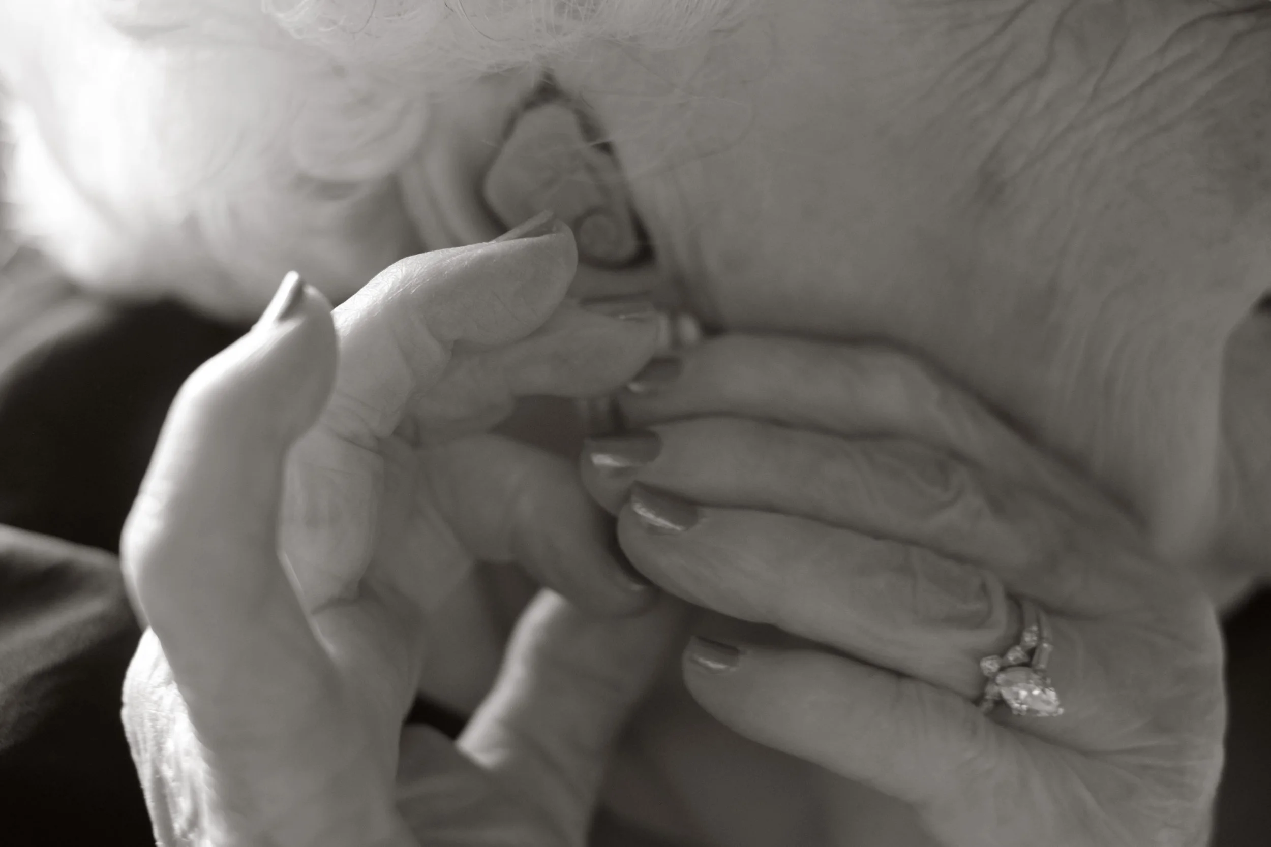 Close-up black-and-white photo of a person's hands gently touching another person's face and chest, with visible jewelry on one finger.