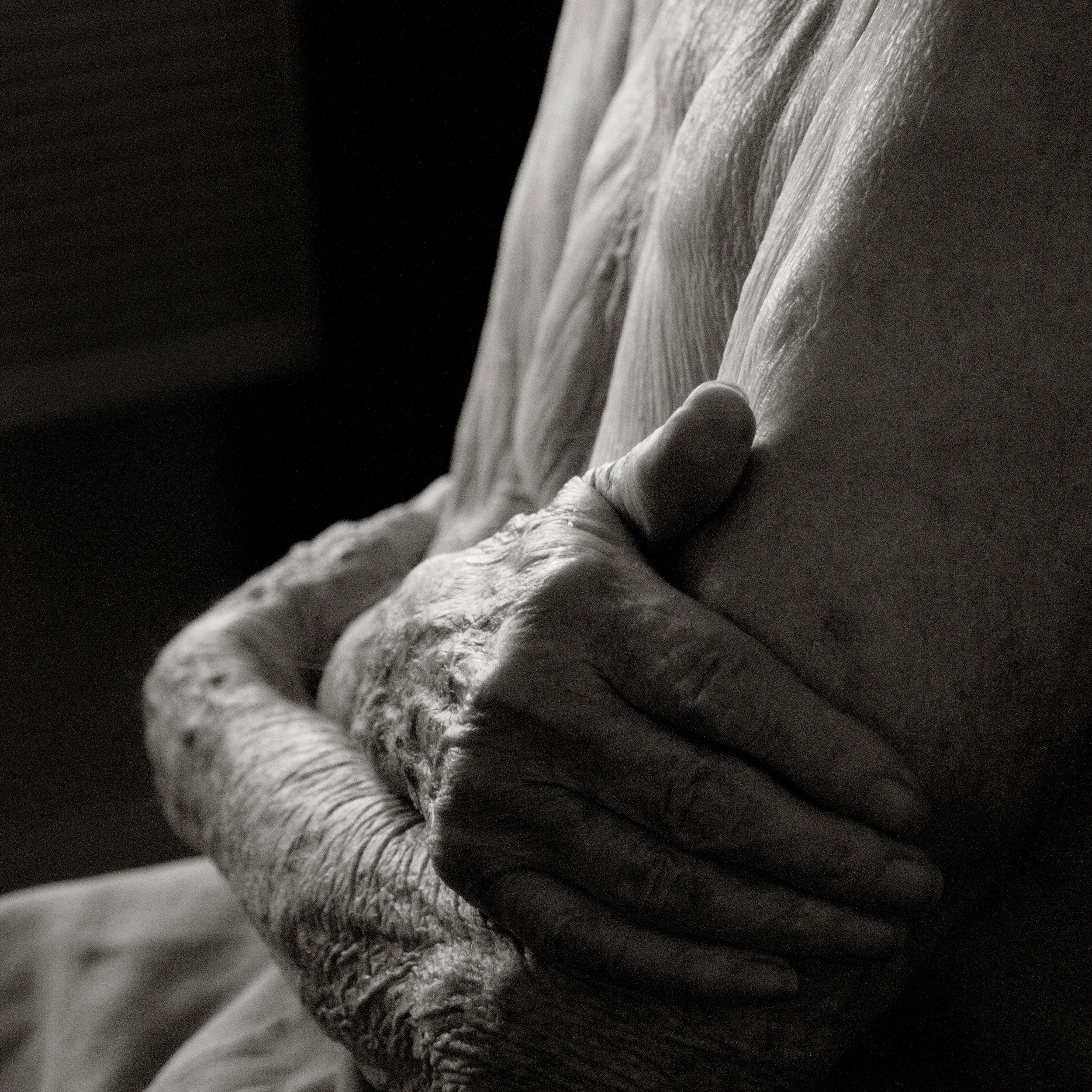 Close-up black-and-white photo of an elderly person's wrinkled hand resting on their upper arm.