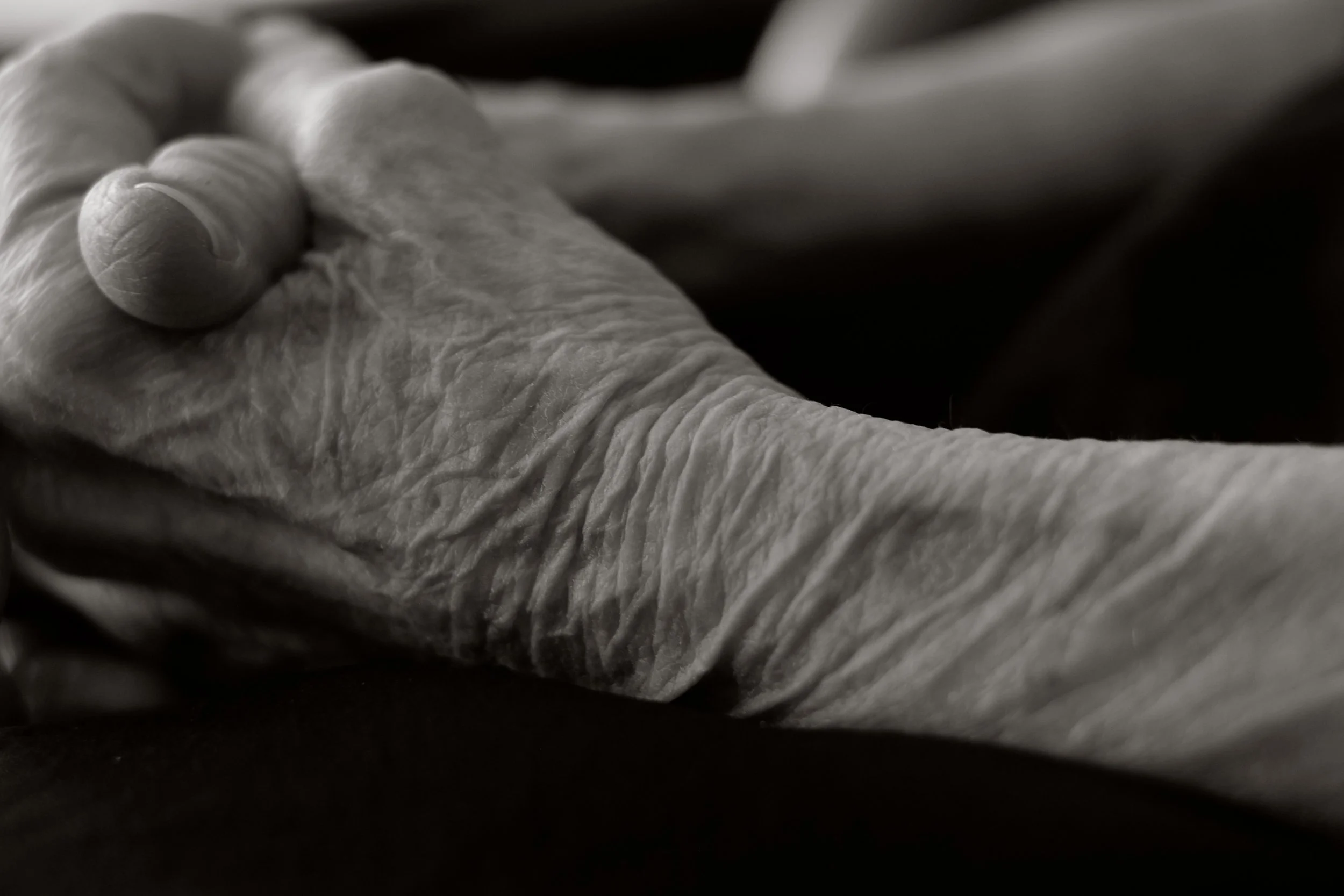 Close-up black and white photo of wrinkled, aged hand with visible veins resting on a dark surface.