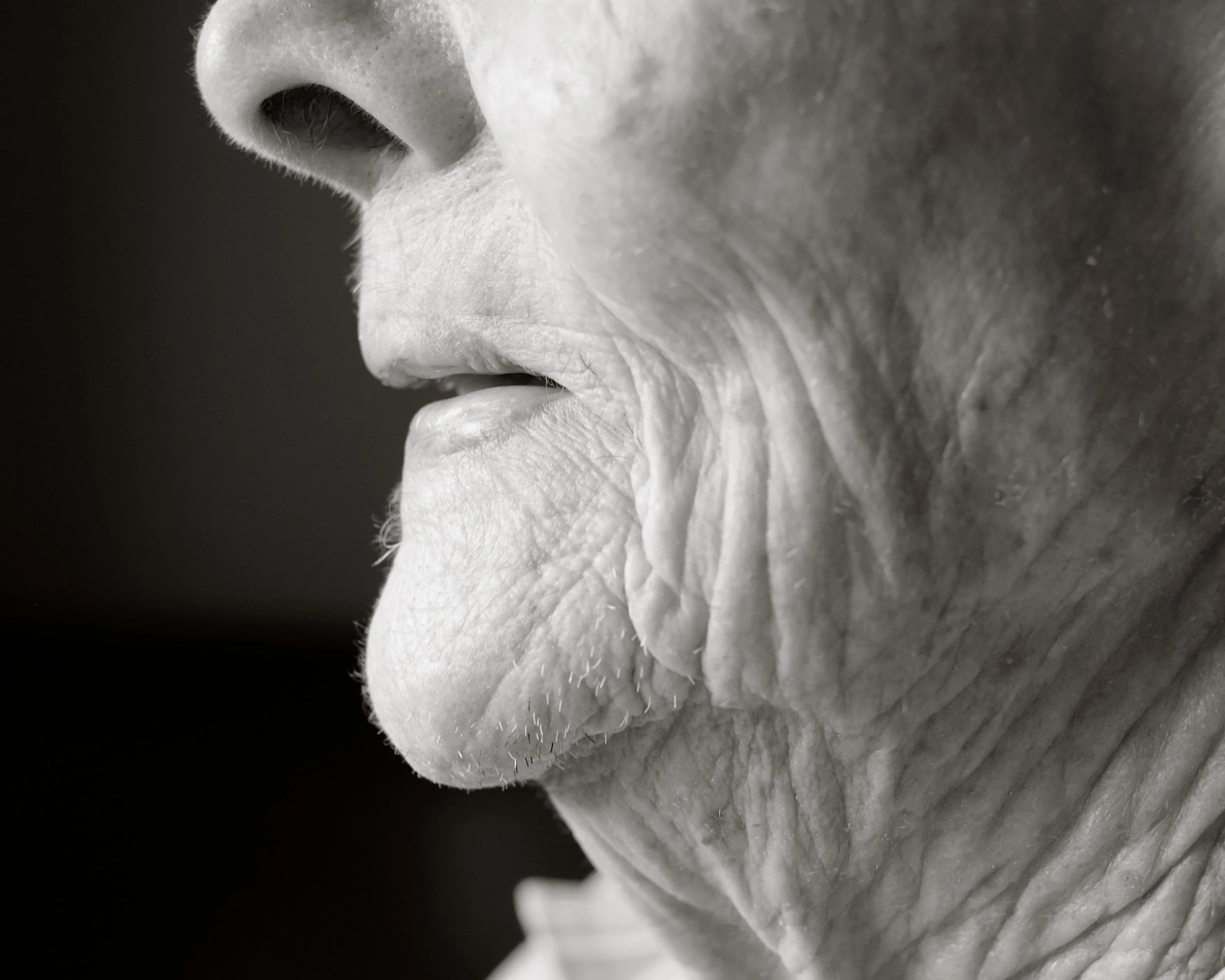 Close-up black and white photo of an elderly person's face, focusing on the nose, lips, chin, and wrinkled skin.