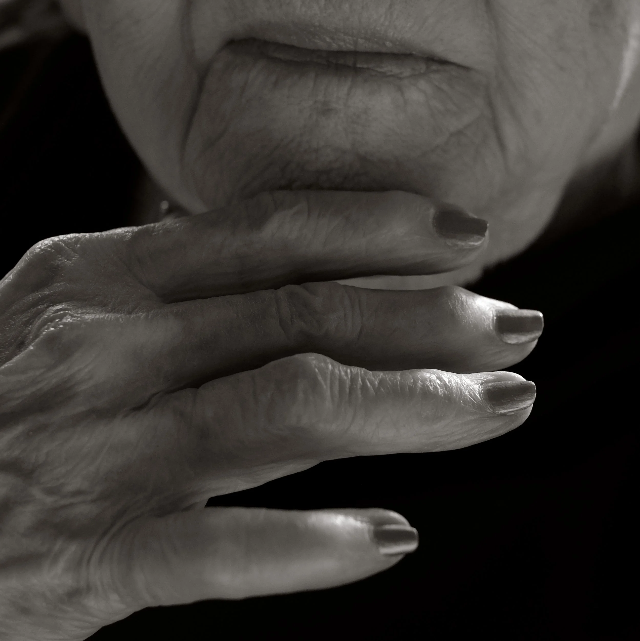 A close-up black and white photograph of an elderly person's face with wrinkled skin and closed lips, with their hand resting on their mouth.