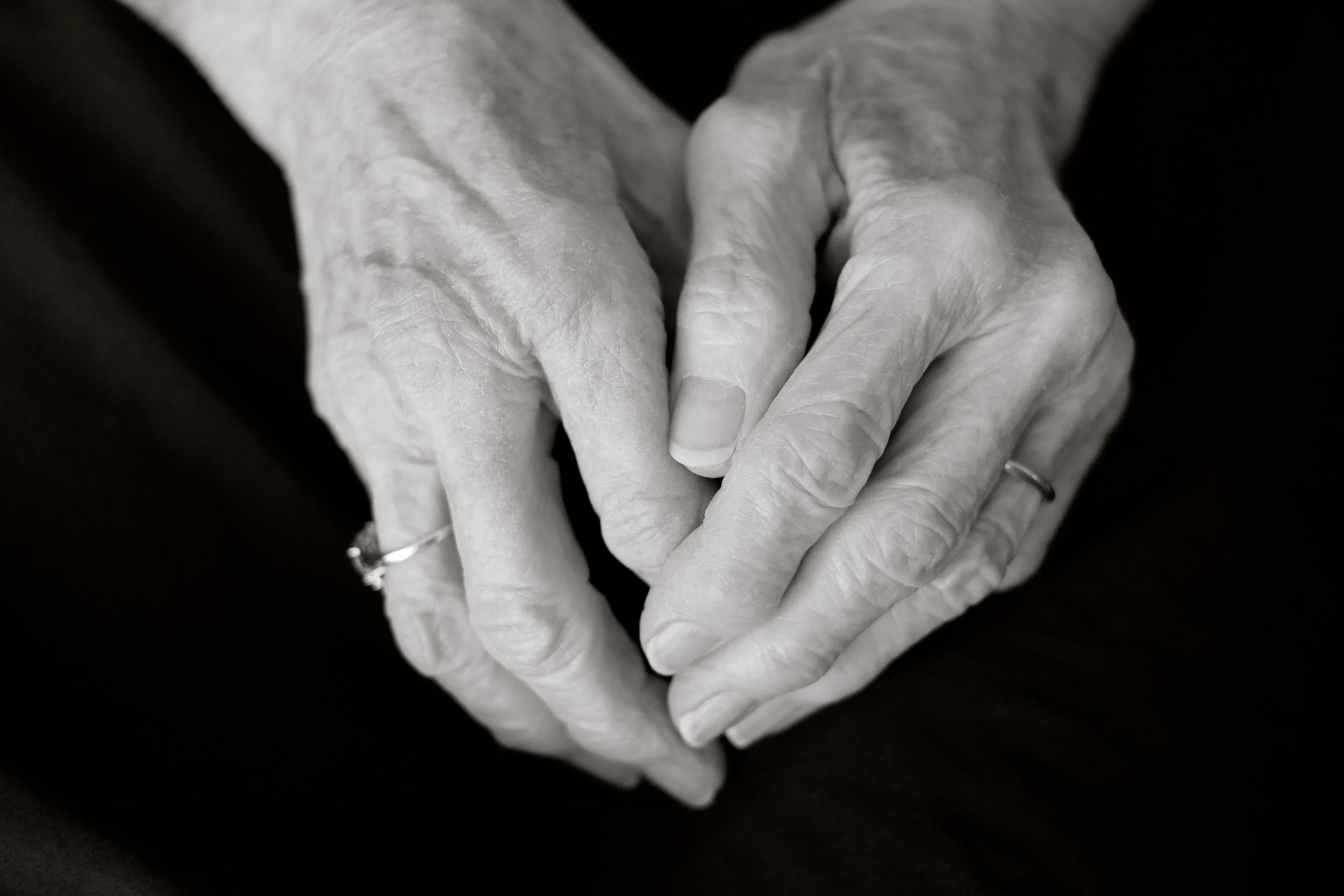 Close-up of elderly hands with visible veins and wrinkles, gently clasped together, on a dark background.