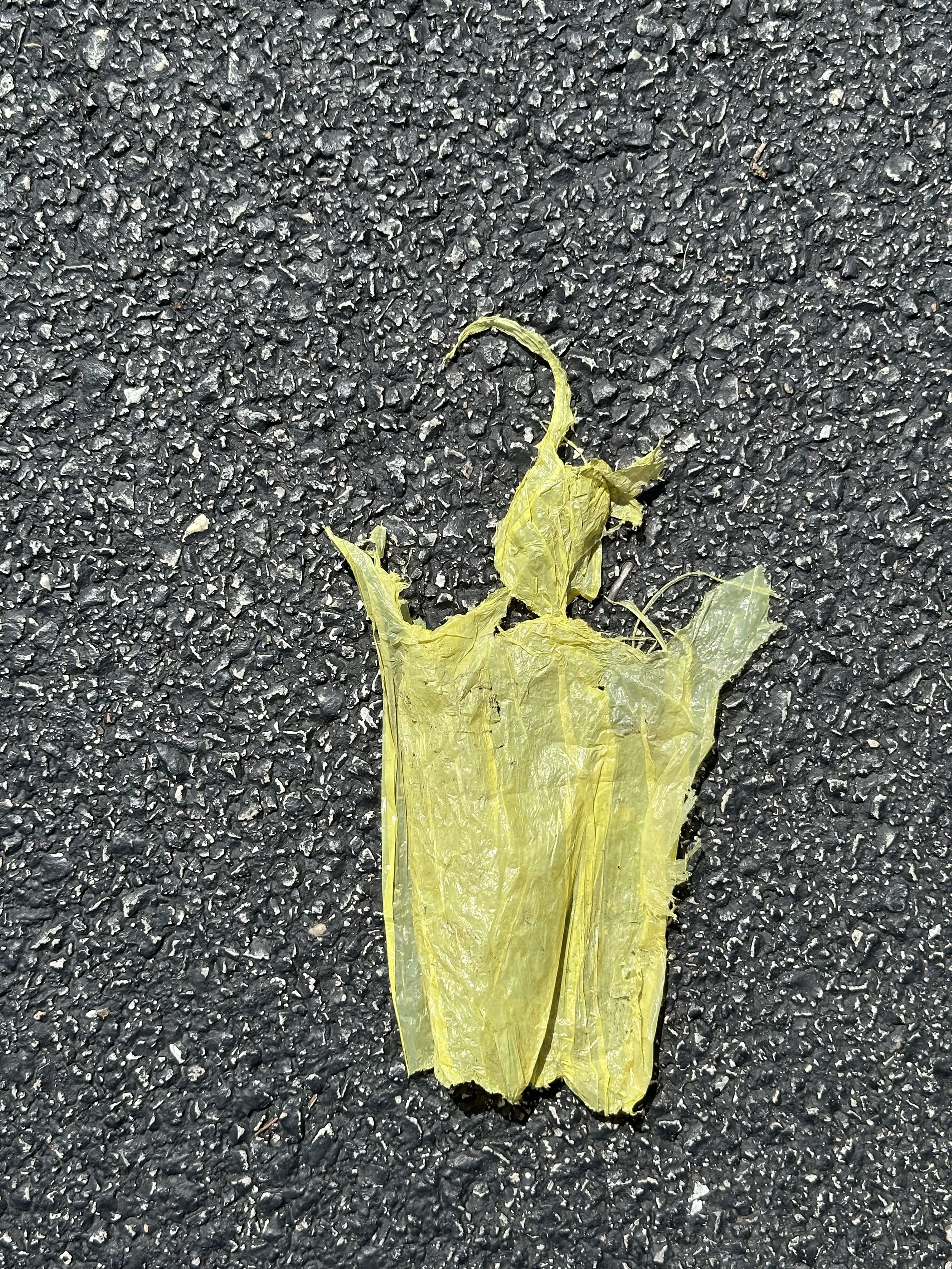 A yellow piece of yellow plastic that resembles a person lies on a rough, black asphalt surface.