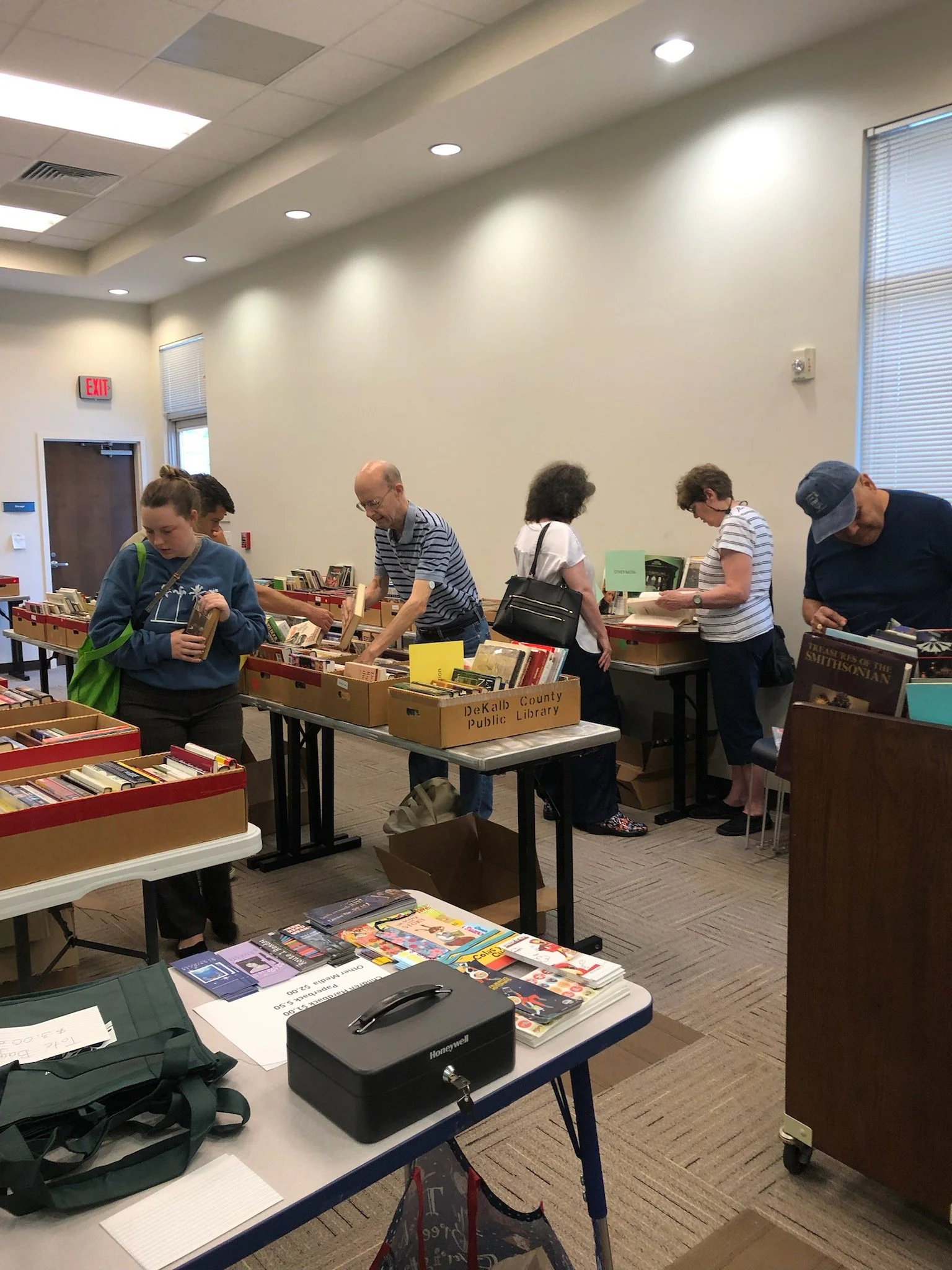 People browsing books at a used book sale organized by DeKalb County Public Library in a room with white walls and large windows with blinds.