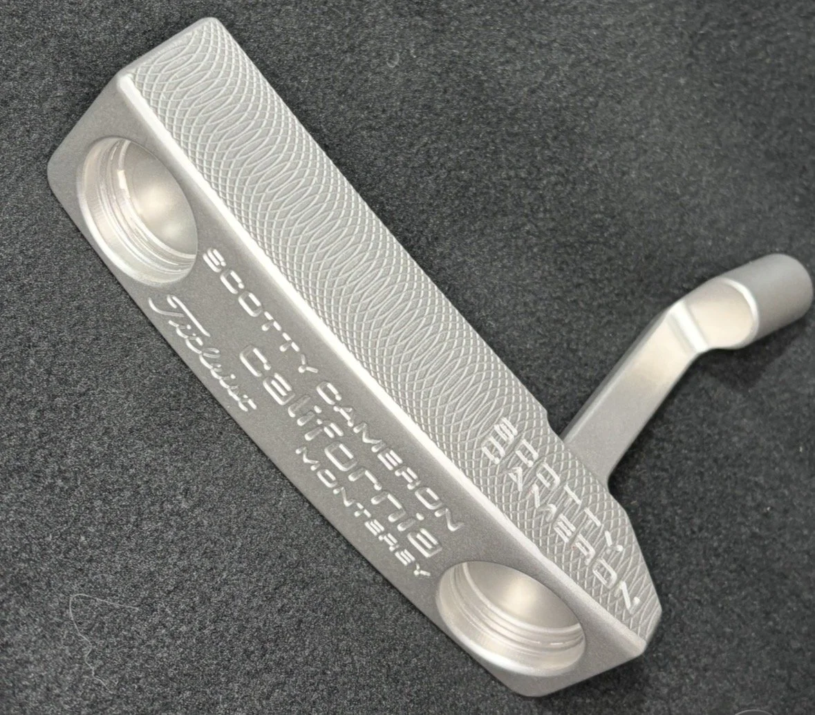 A close-up of a silver CNC-machined keychain shaped like a putter golf club, with engraved text and detailed grip pattern, resting on a black textured surface.