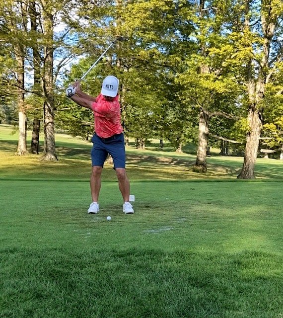 A man taking a golf swing on a golf course surrounded by trees, wearing a cap, red shirt, and blue shorts.
