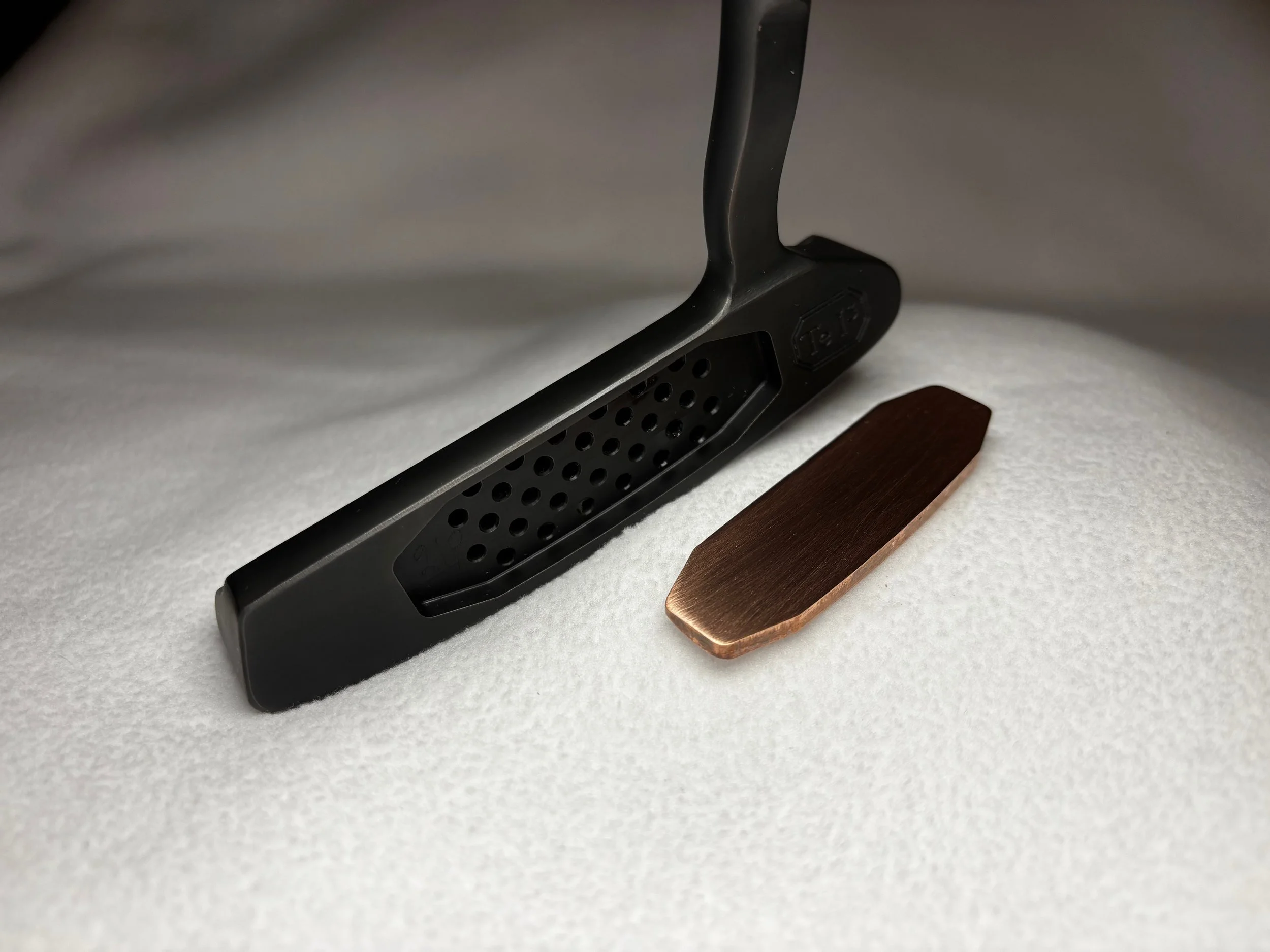 Close-up of a black, perforated metal object with a label, resting on a small wooden stand on a white surface.