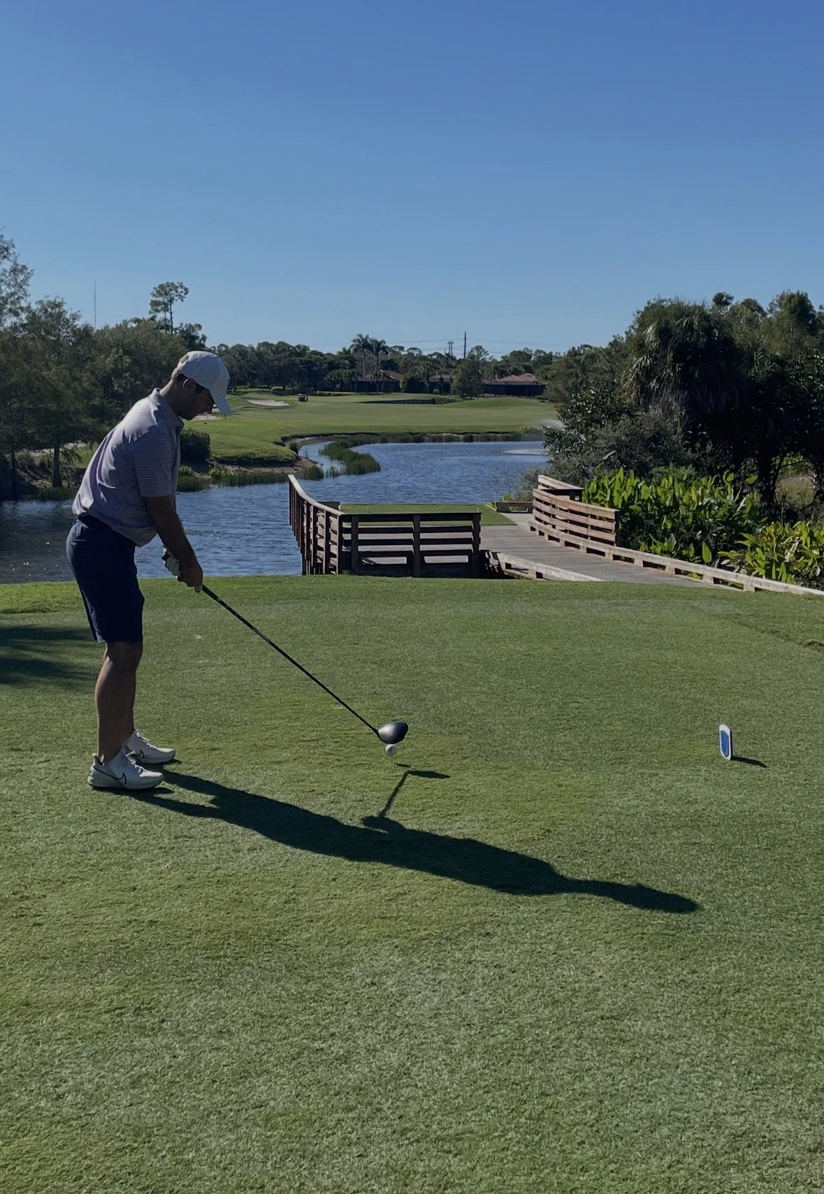 A person standing on a golf course preparing to hit a golf ball, near a body of water and a bridge, with trees and a clear blue sky in the background.