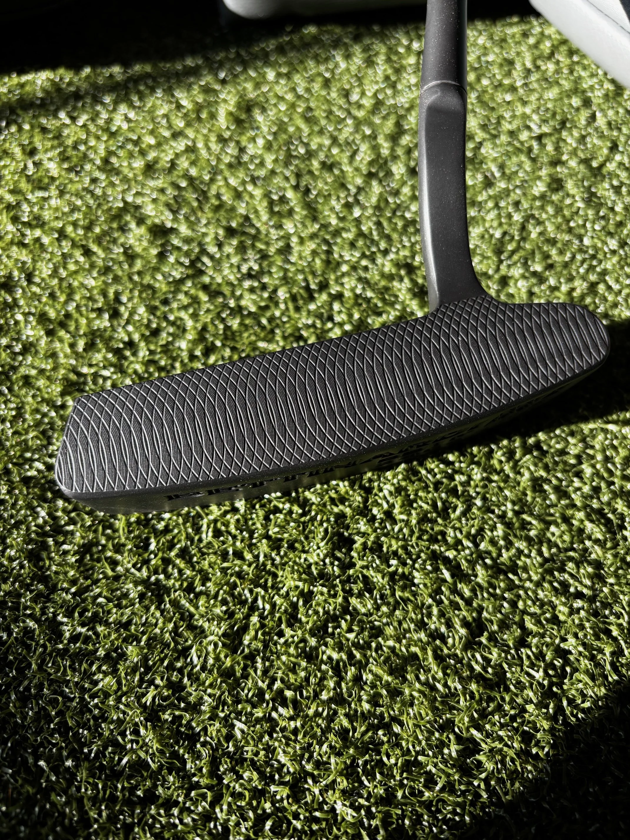 Close-up of a golf putter lying on artificial grass turf, showing the textured face of the club.
