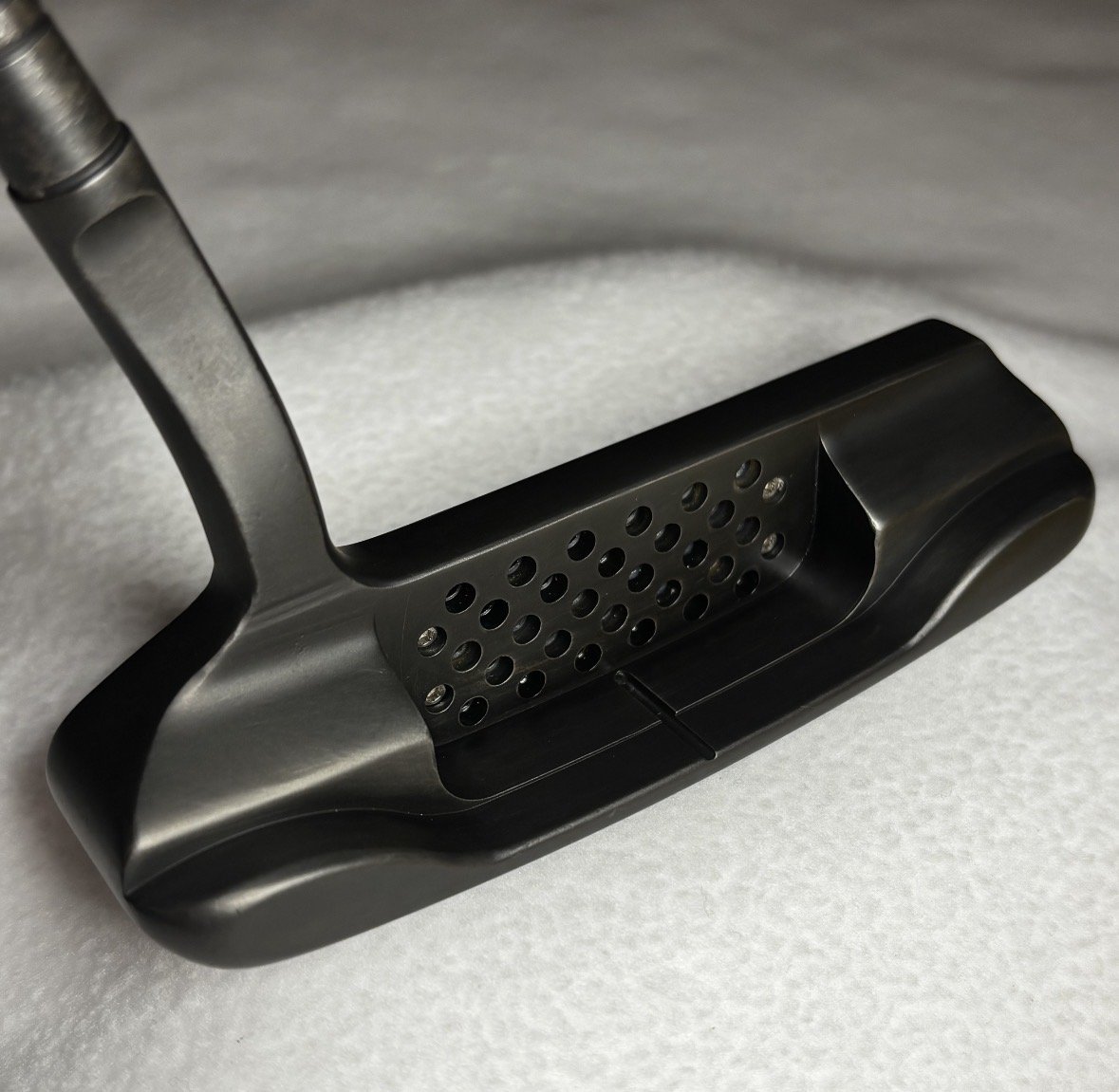 A black golf putter with a perforated face and a curved sole, resting on a light surface.
