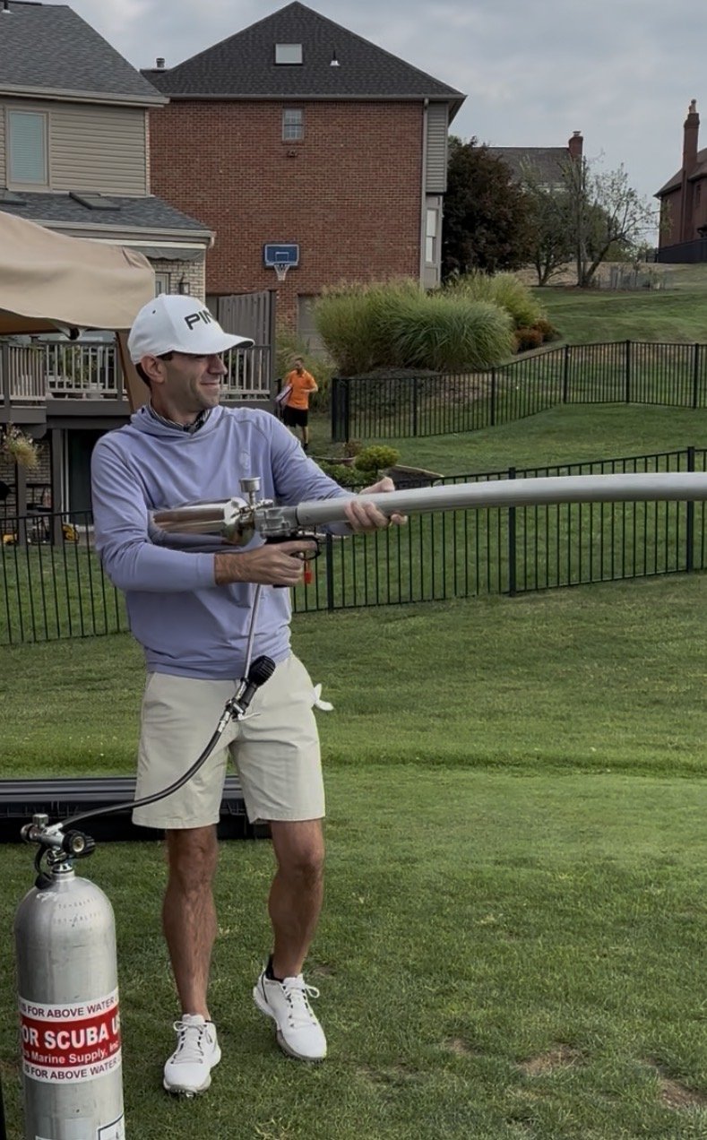 A man in a white cap and purple long-sleeve shirt holding and aiming an air gun with a long barrel, connected to a scuba tank, on a grassy backyard with houses in the background.