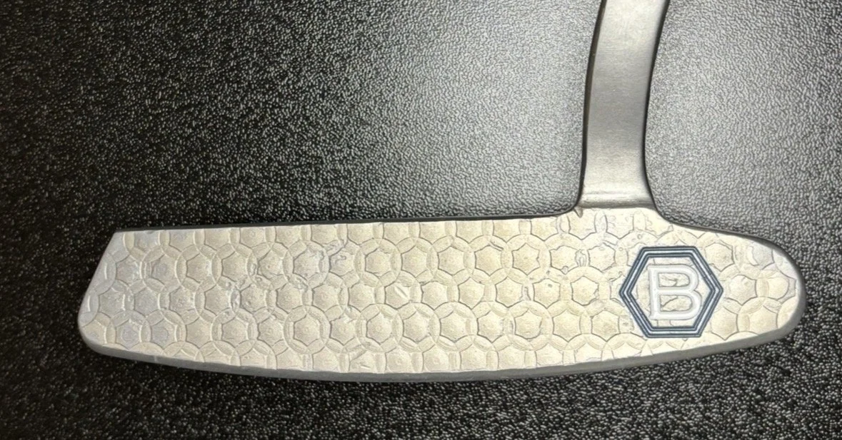 Close-up of the heel of a golf club with a textured white and gray design and a capital letter 'B' inside a hexagon on the back of the club.