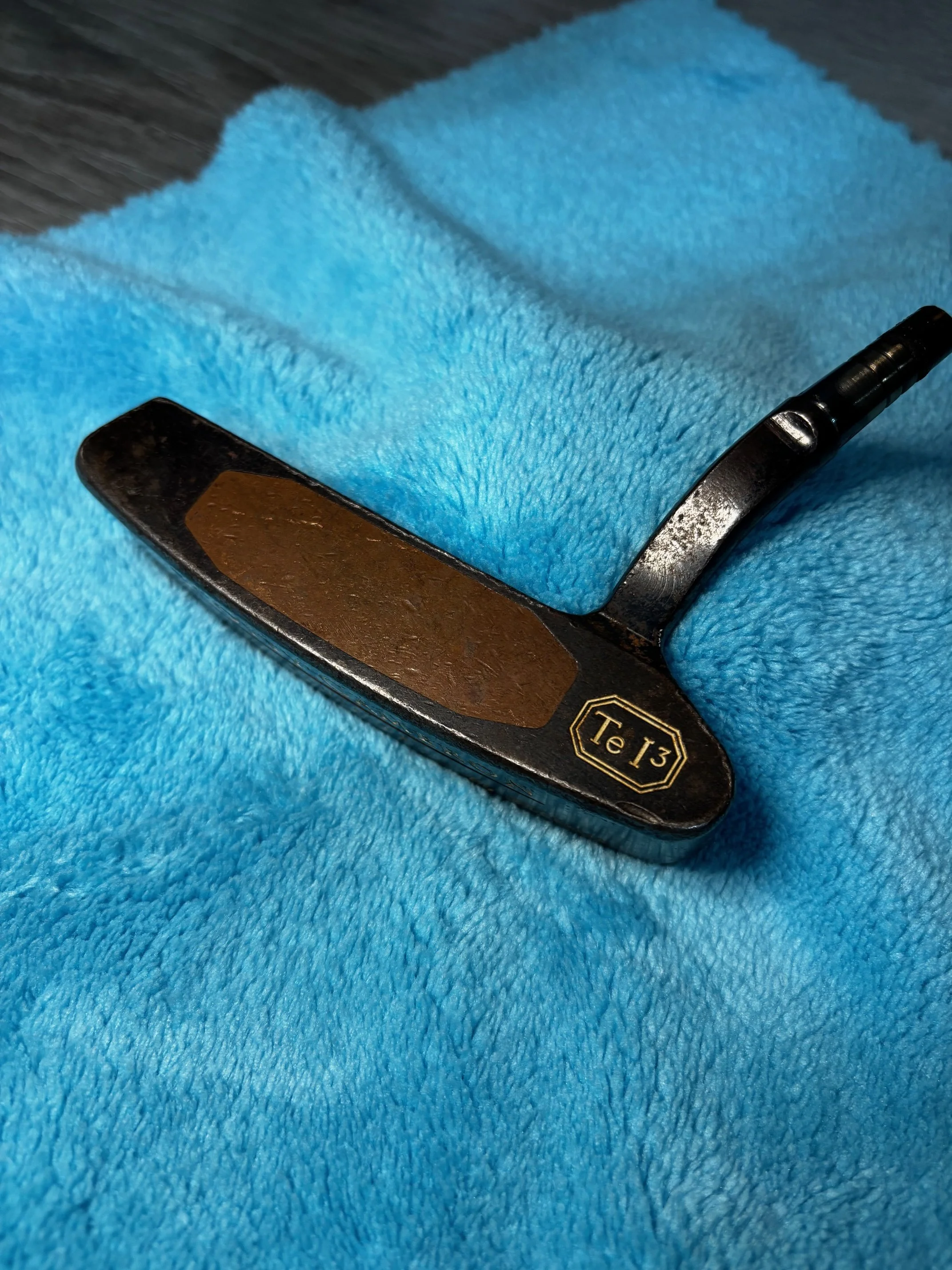 Close-up photo of a straight razor with a black handle and a gold label marked 'Te 13,' resting on a bright blue fluffy fabric surface.