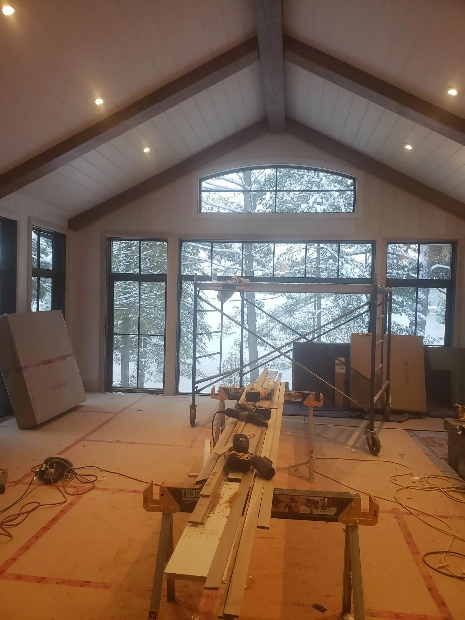 Interior of a house under construction with large windows, construction tools on a workbench, and scaffolding in a snowy landscape outside.