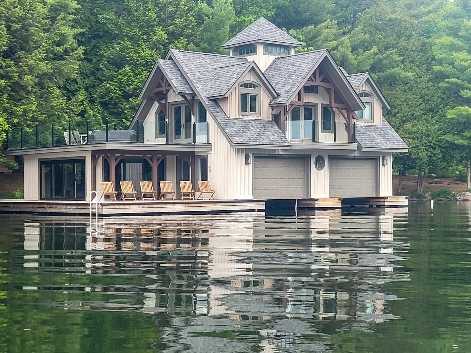 A modern house built on a lake with a reflecting water surface. The house has multiple gabled roofs, large windows, and a deck with seating. The background features lush green trees on a hillside.