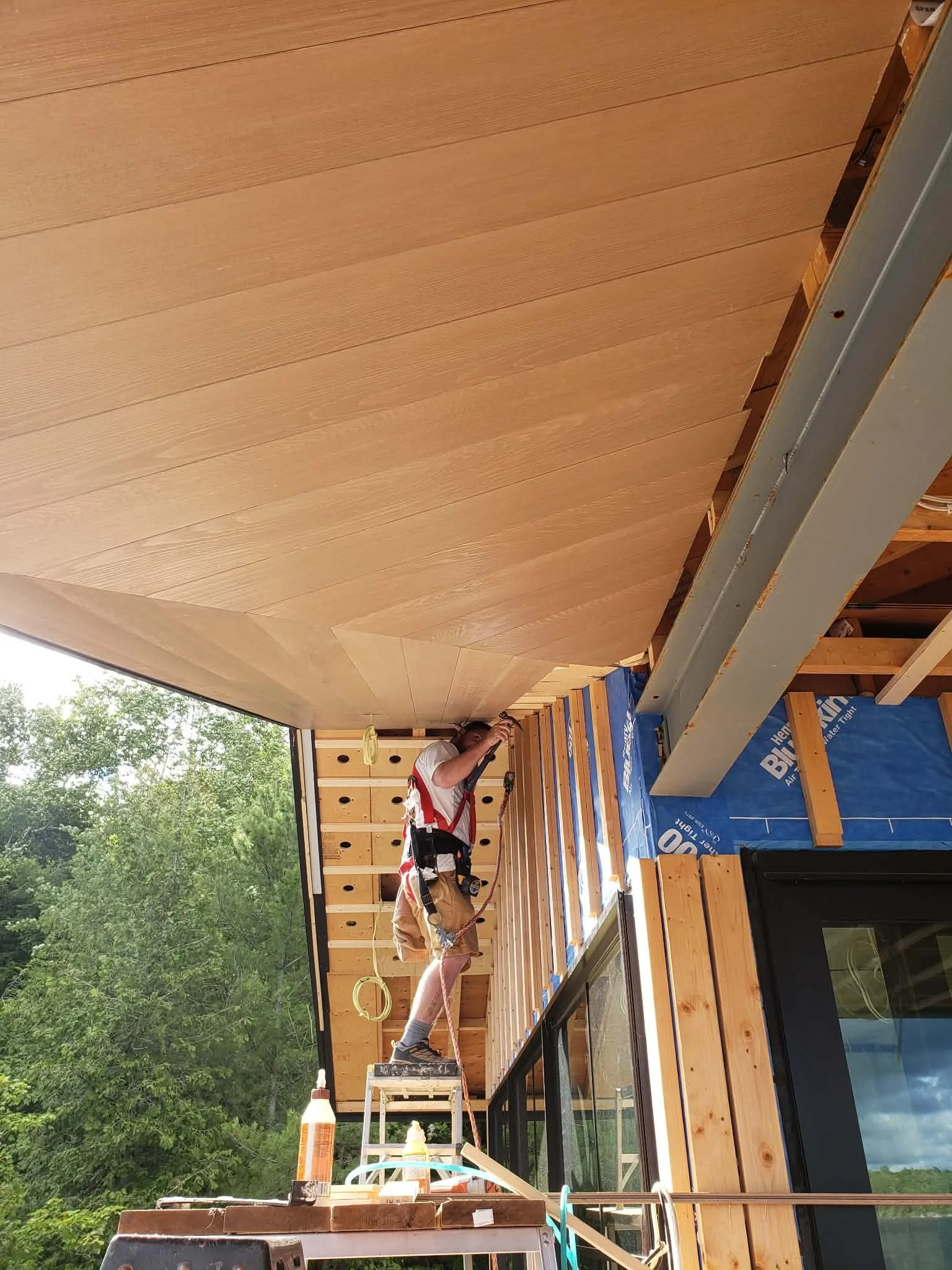 A construction worker installing wood ceiling panels in a partially built room with large windows and a view of green trees outdoors.