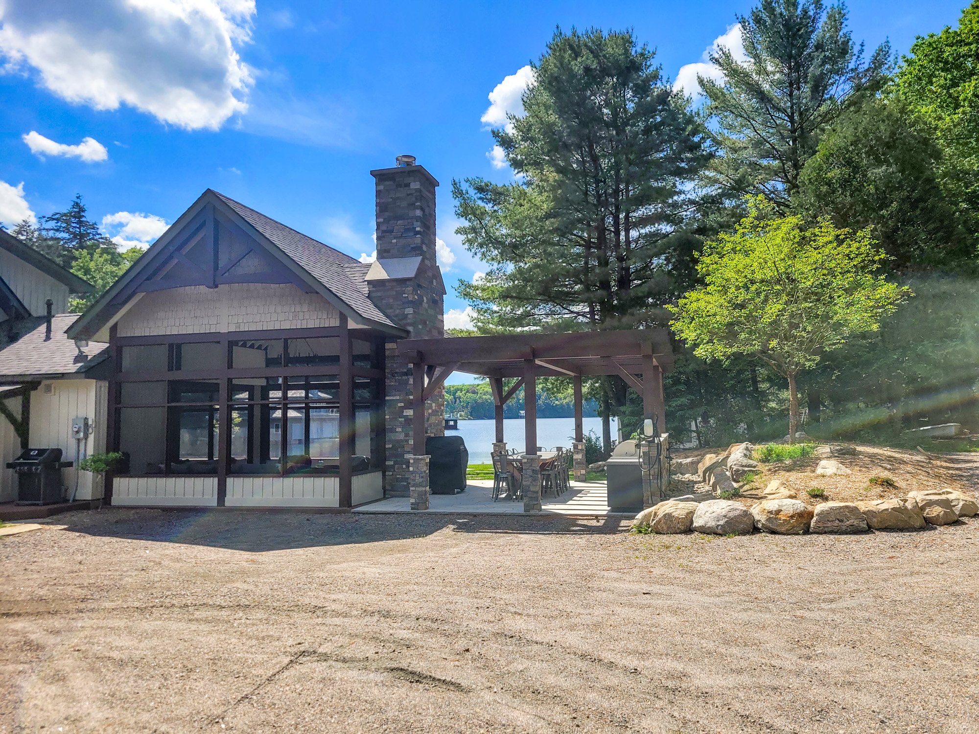 A house with a stone chimney, screened porch, and outdoor kitchen area near a lake, surrounded by trees under a blue sky with clouds.