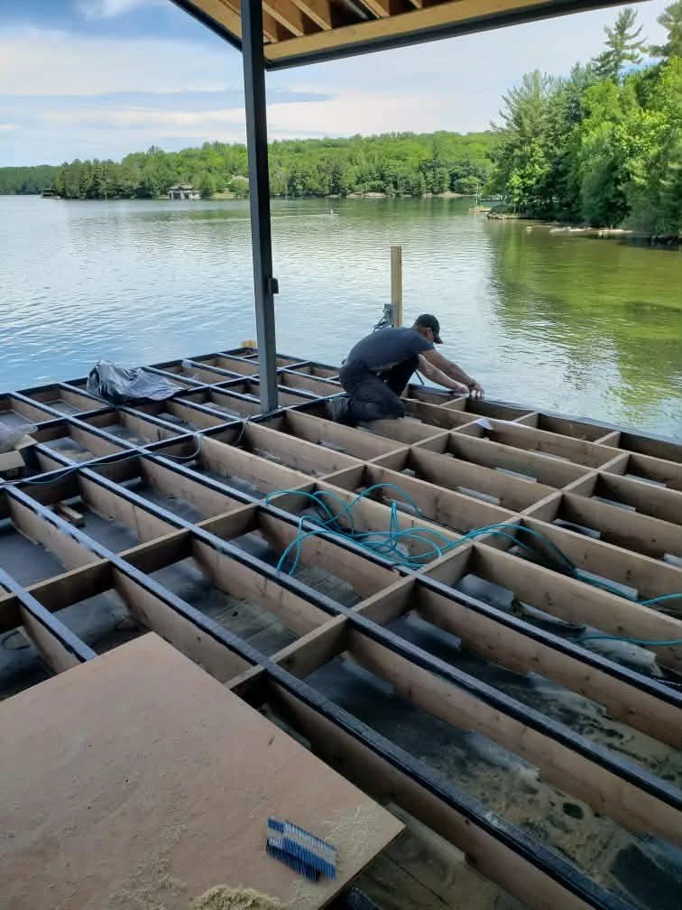 A man working on the wooden framework of a house built over a body of water, with trees and a blue sky in the background.